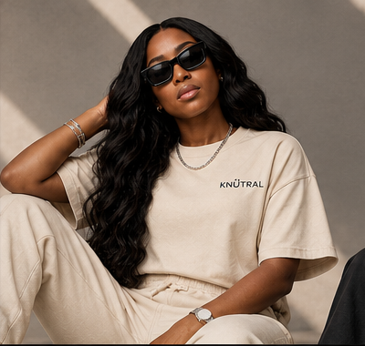 Stylish woman sitting, wearing sunglasses and a beige T-shirt with the word "KNUTRAL" on it, against a neutral background.