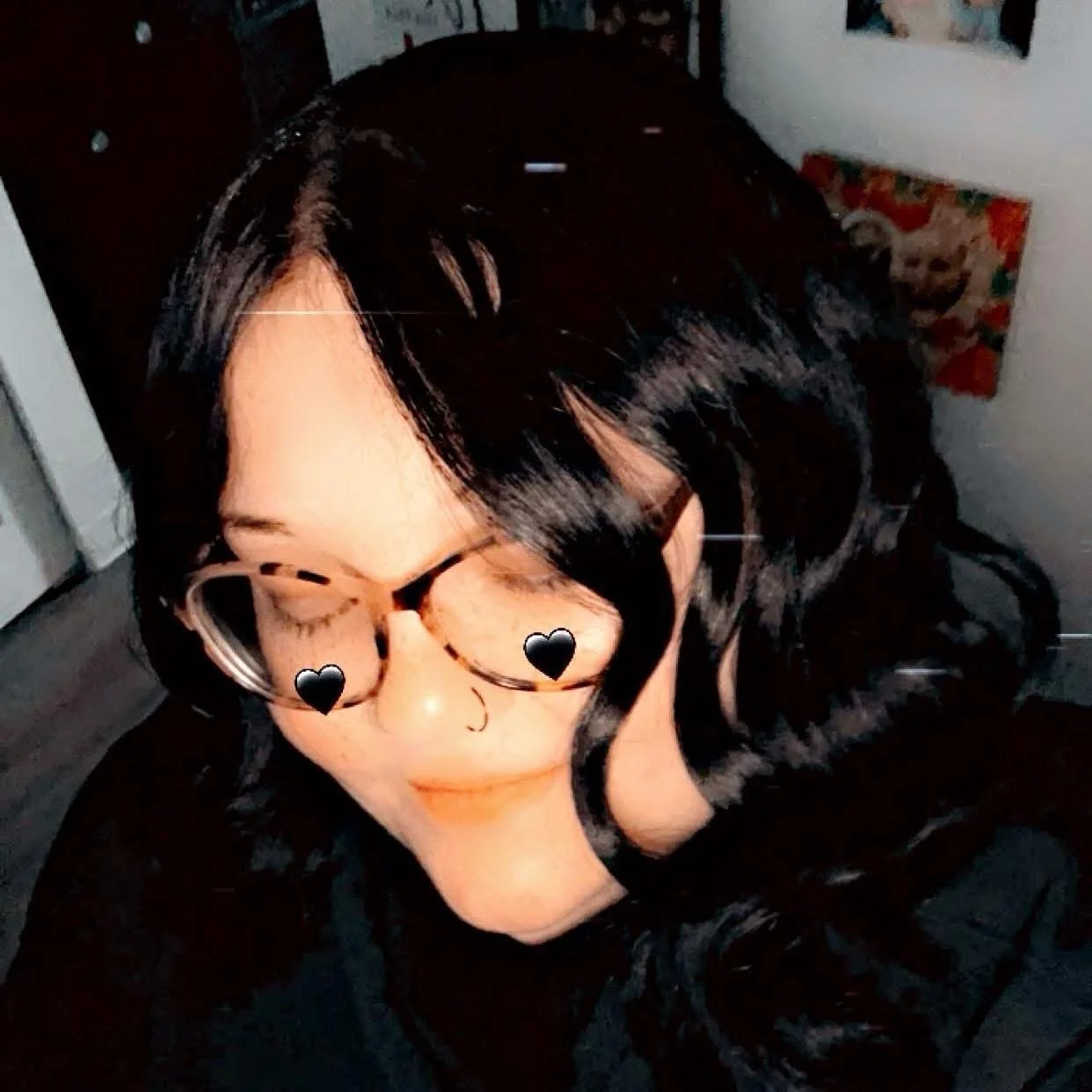 Close-up of a woman with black hair, wearing glasses, with emoji overlayed on her face, and eyes closed. She appears to be smiling slightly. The background shows parts of a room with posters on the wall.