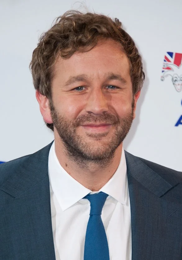 A man with light brown, curly hair and a beard wearing a dark suit, white shirt, and blue tie, smiling in front of a backdrop with logos.