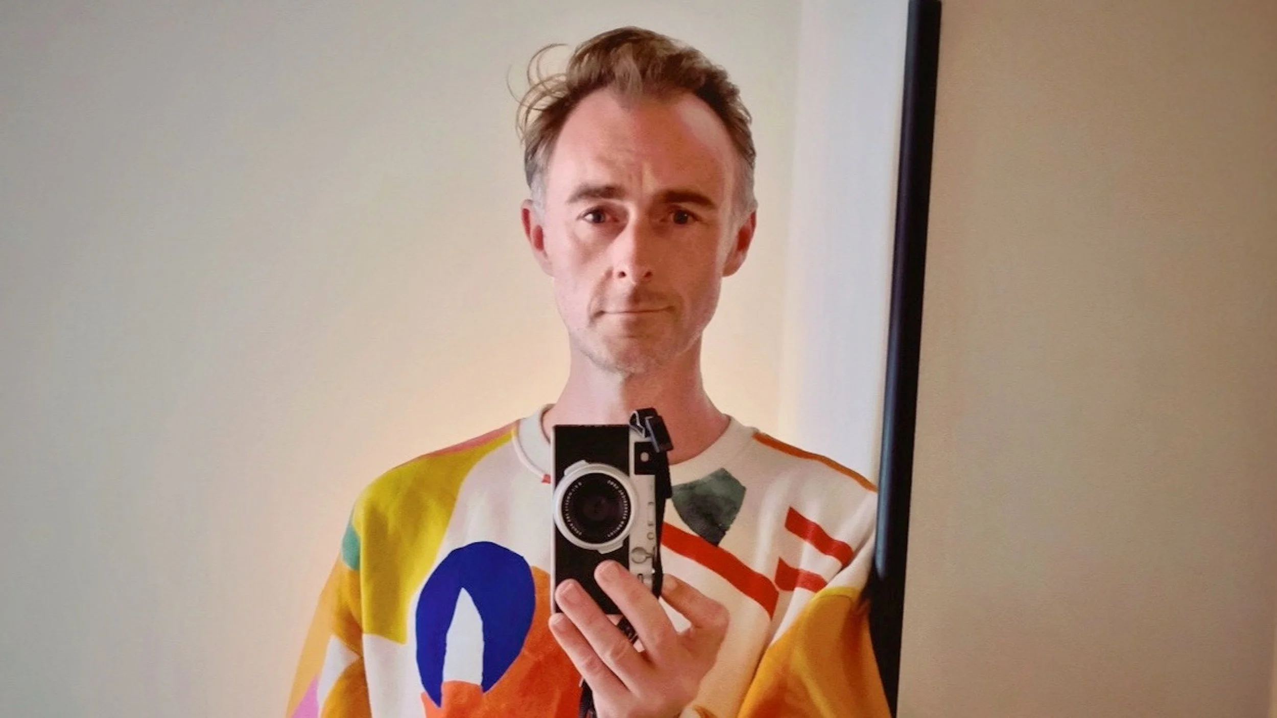 Person taking a selfie in front of a mirror, holding a camera, wearing a colorful shirt with geometric patterns, with a plain wall background.