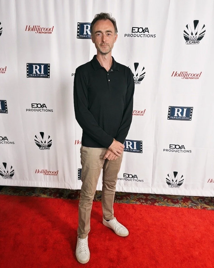 A man standing on a red carpet in front of a white backdrop with logos, wearing a black long-sleeve polo shirt, khaki pants, and white sneakers.