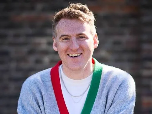 Young man with light brown hair smiling outdoors, wearing a gray cardigan with red and green accents over a white shirt.