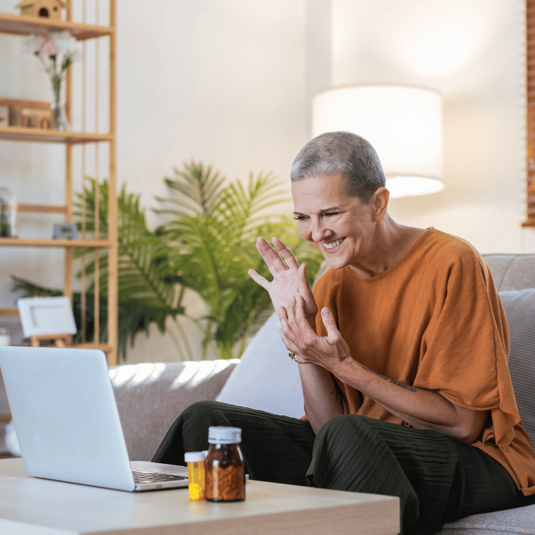 A woman with short gray hair smiling and waving at a laptop while sitting on a couch in a living room, with medication bottles on the table in front of her.