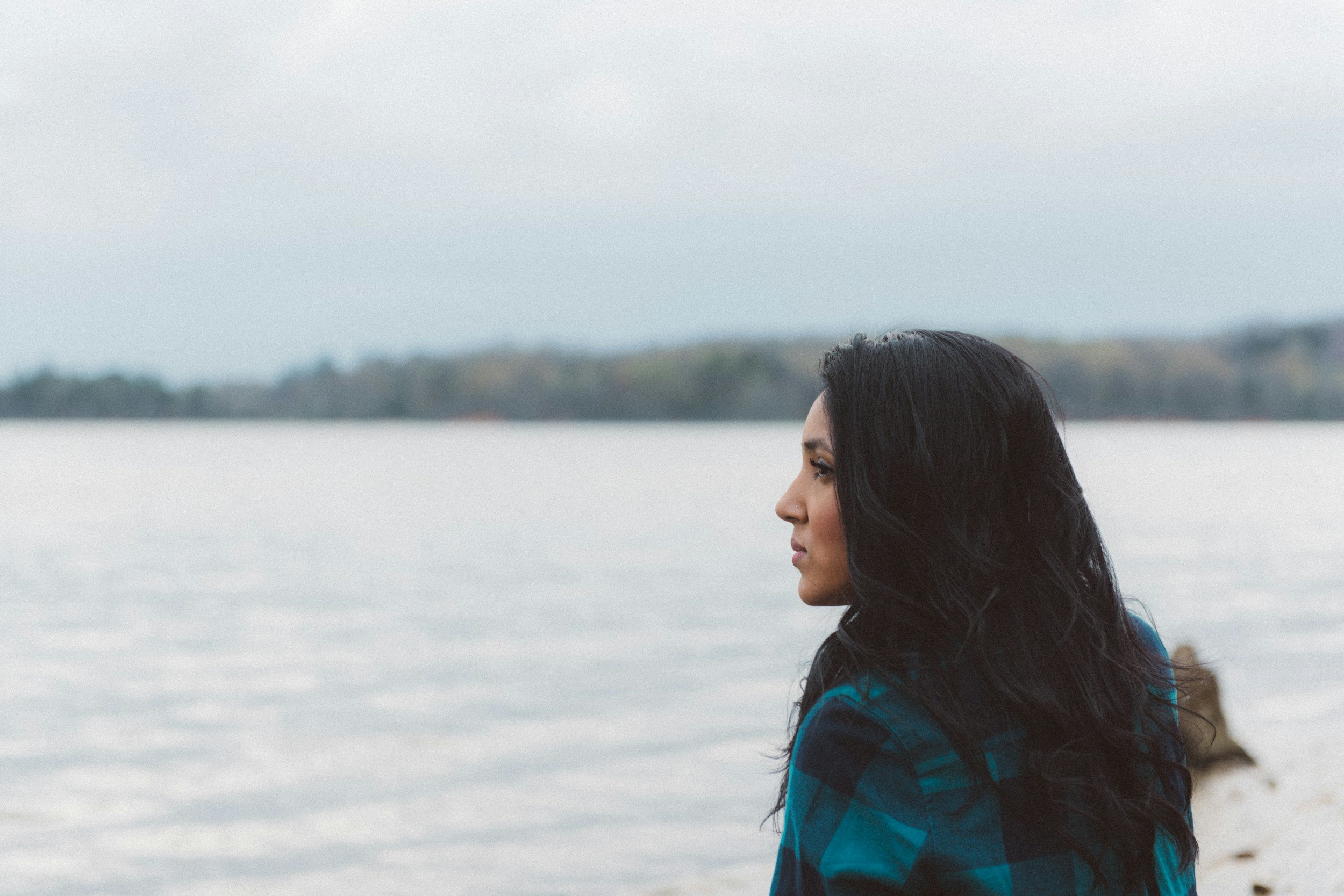 A woman with long dark hair looking out over a body of water, with a distant shoreline and trees in the background.