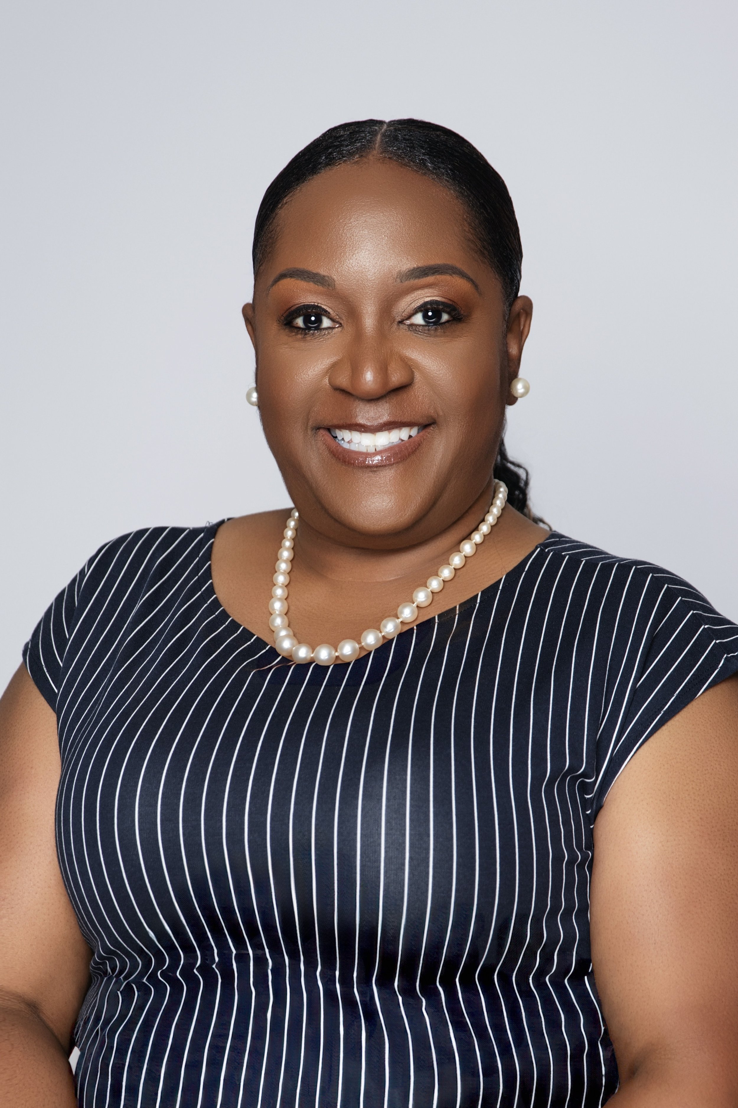 A headshot of a smiling Black woman with dark hair styled back, wearing pearl earrings, a pearl necklace, and a navy blue dress with white pinstripes.
