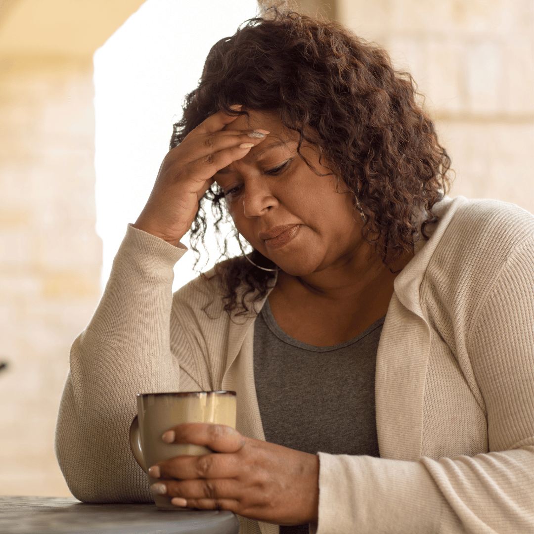 A woman sitting at a table holding a mug, with a worried or distressed expression, resting her head in her hand.