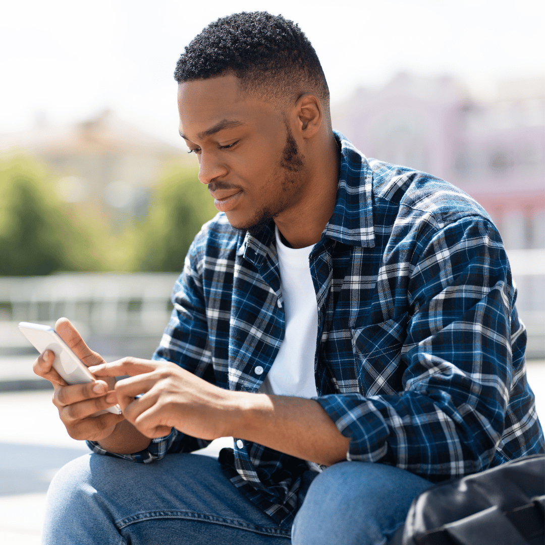A young man in a plaid shirt and jeans sitting outdoors, looking at his smartphone.