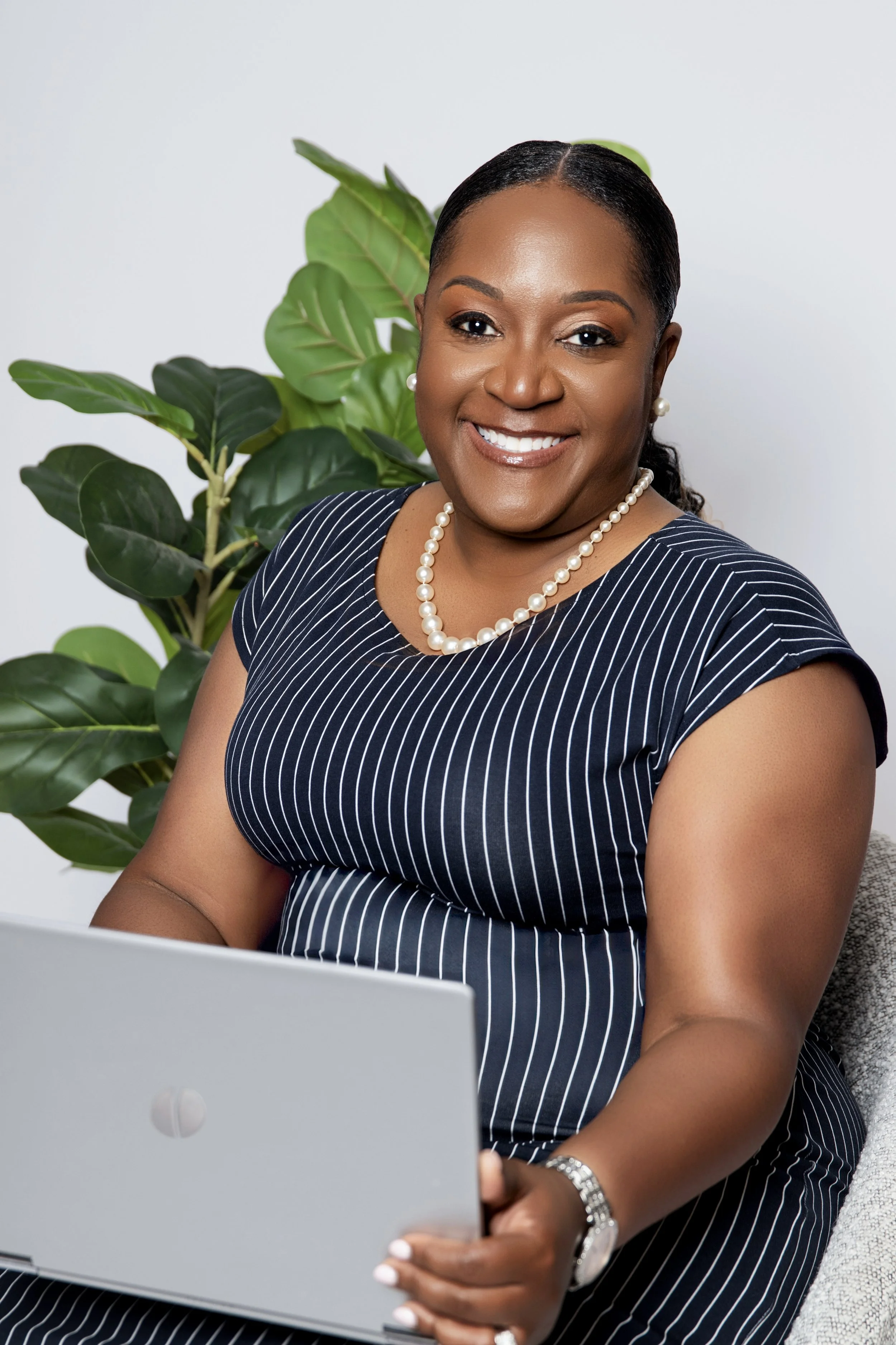 A woman in a navy and white pinstripe dress with pearl jewelry is sitting and looking at a silver laptop, with a large green plant behind her.