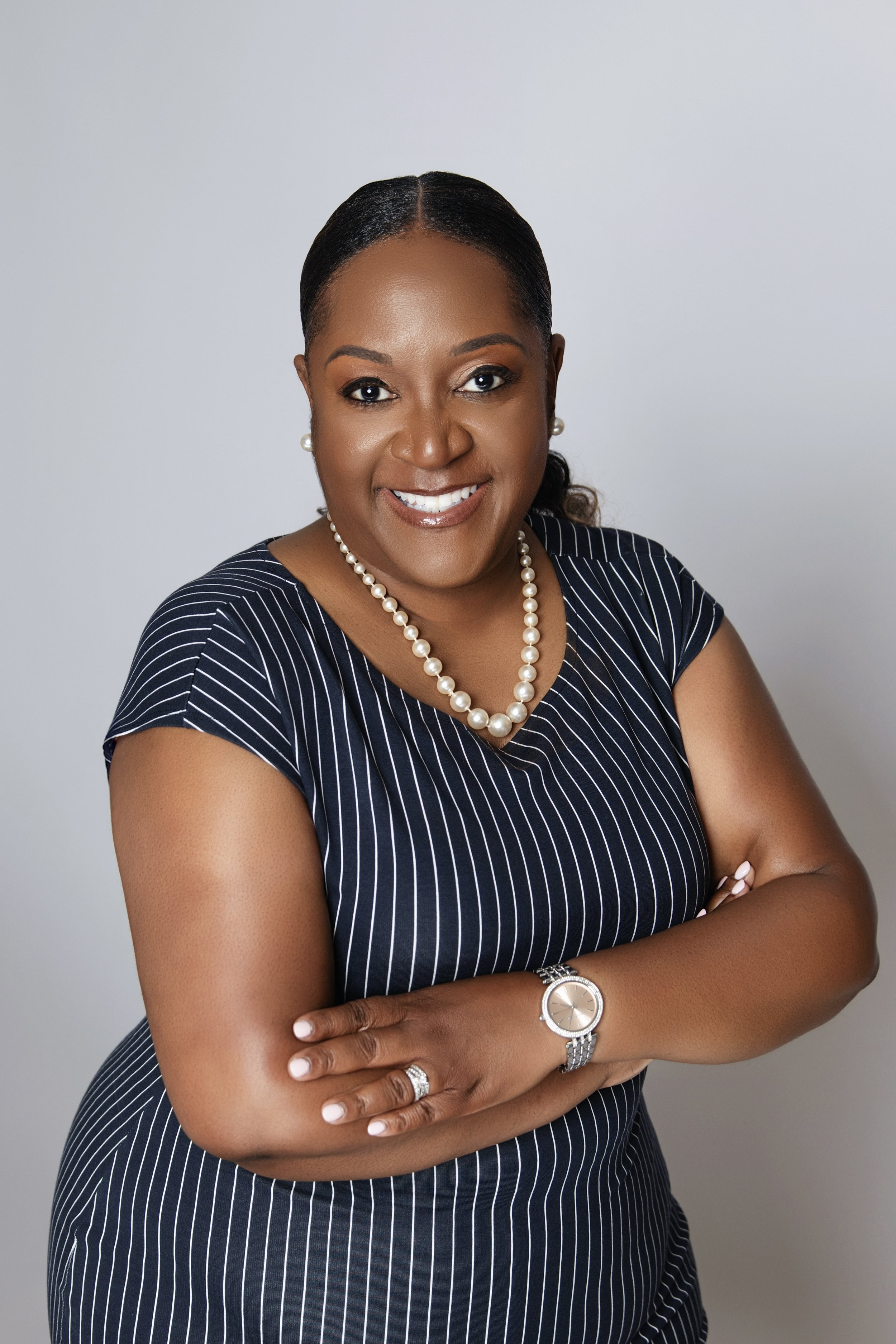 Portrait of a smiling African American woman wearing a navy blue pinstripe dress, pearl necklace, pearl earrings, watch, and ring, with her arms crossed against a plain gray background.