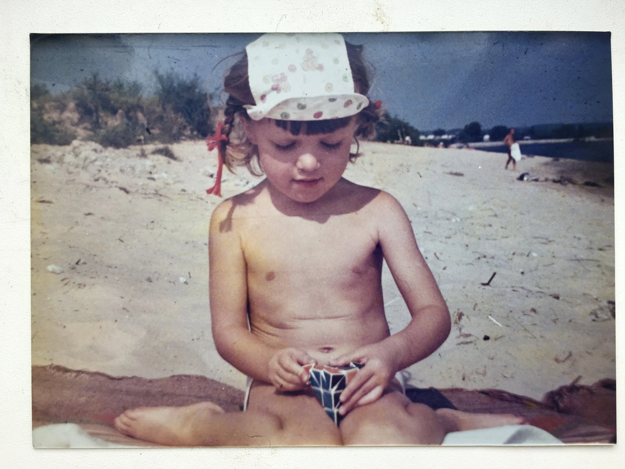 A young child with curly hair, wearing a polka dot sun hat, sitting on a beach, playing with a small puzzle toy.