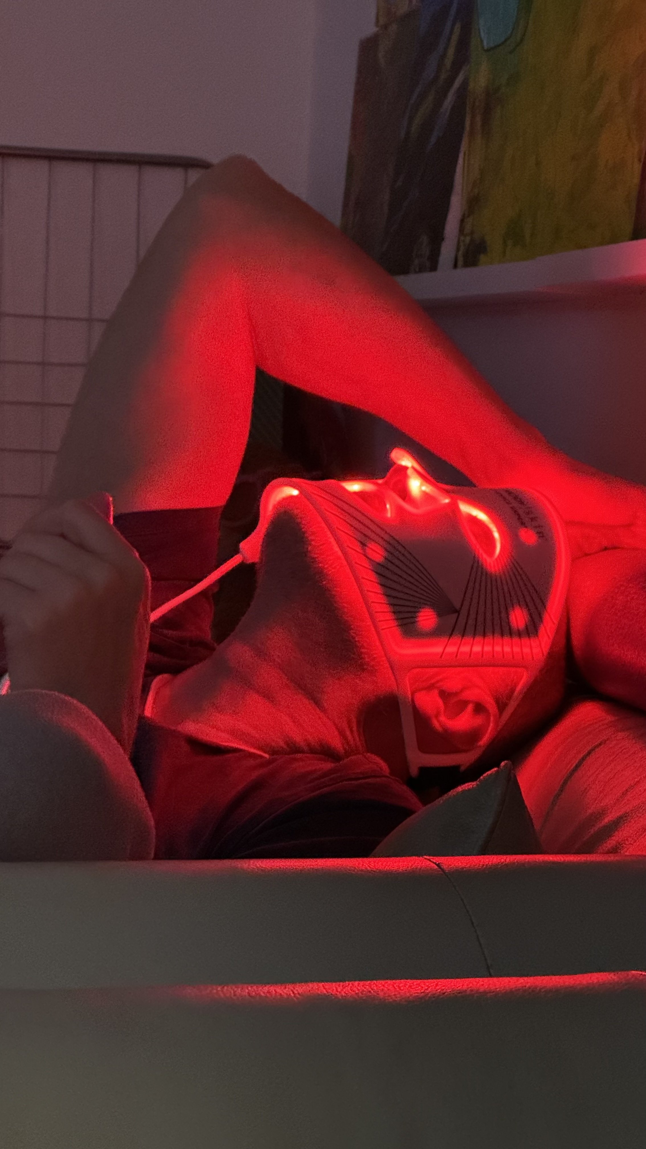 Person lying down wearing a hoodie with a red LED light projecting onto their face and body, holding an electronic device.