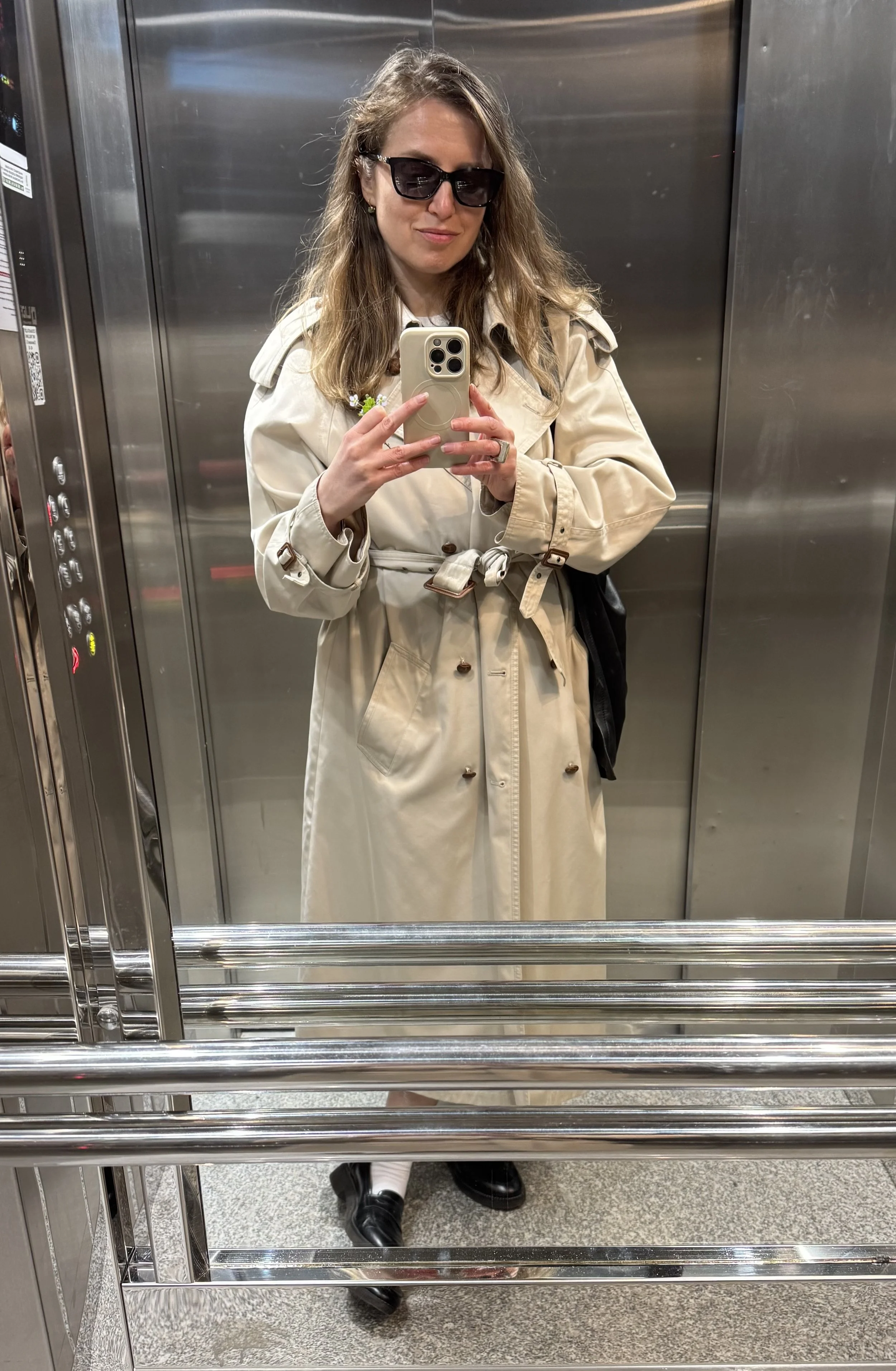 A woman with wavy brown hair, wearing black sunglasses and a beige trench coat, taking a selfie in an elevator mirror. She holds a striped black and white bag, wears black boots, and has a small flower tucked in her coat pocket.