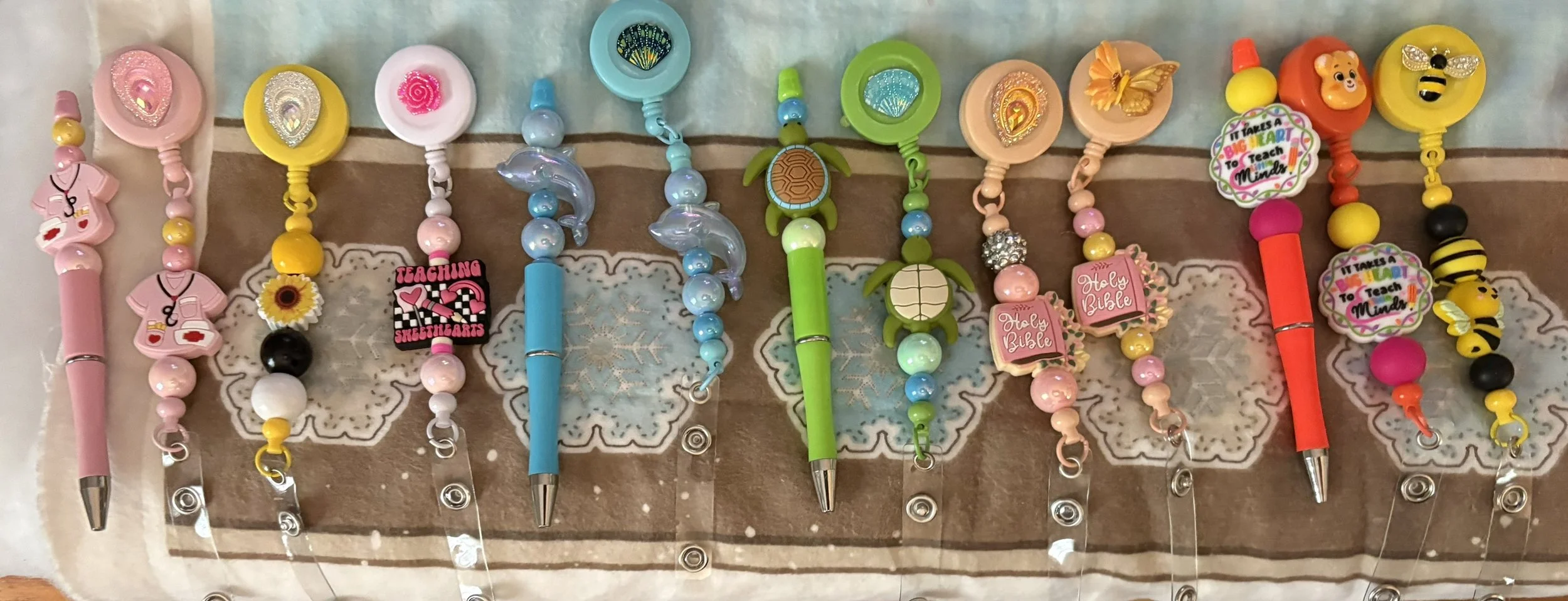 A display of colorful pens, stylus, badge reels, keychains and bookmarks decorated with various themed charms and beads, including animals, hearts, flowers, and inspirational messages. Available in coordinating sets or sold separately.