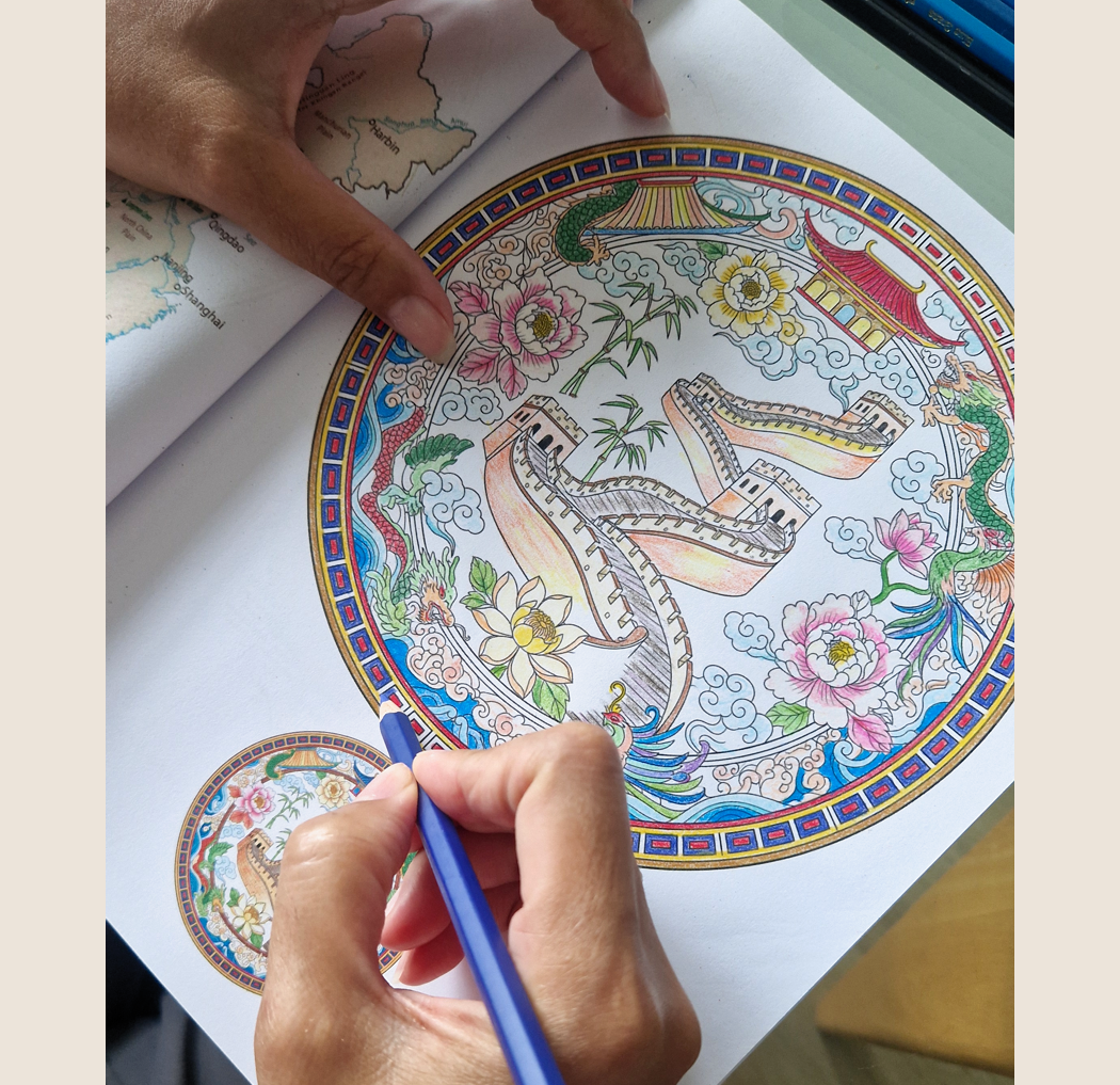 A person coloring a circular picture featuring the Great Wall of China, flowers, clouds, and traditional Chinese architecture. The person is using a blue colored pencil, and the image shows the person's hand partially as they add color to the scene.