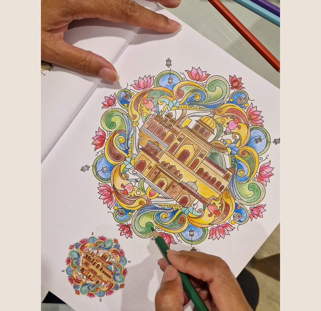 A person's hands coloring a detailed, colorful illustration of a castle surrounded by floral and scroll patterns on a sheet of paper, with a smaller version of the same design nearby.