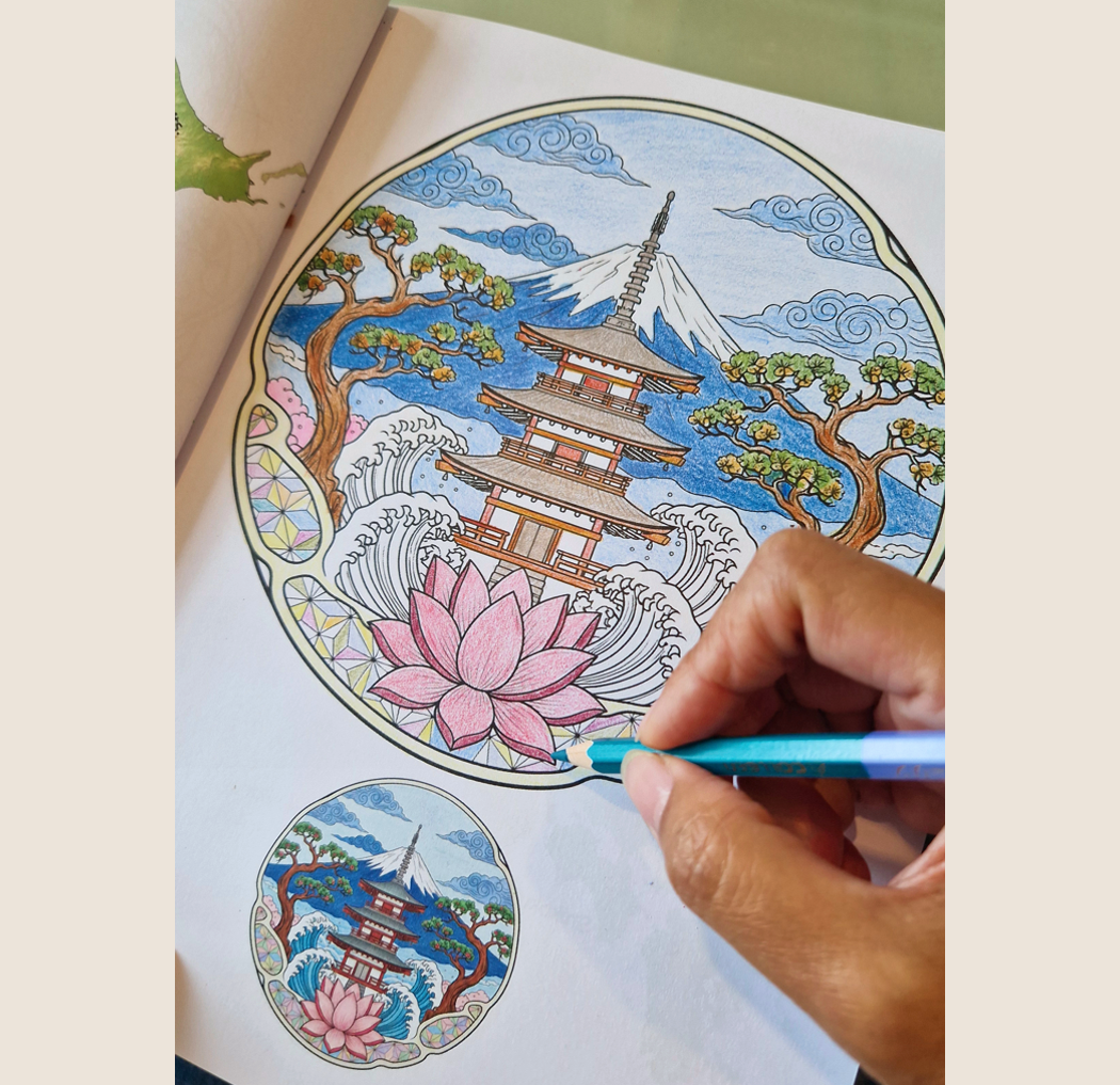 A person coloring a detailed circular illustration of a Japanese pagoda, cherry blossom, Mount Fuji, pine trees, waves, clouds, and a large pink lotus flower with colored pencils.