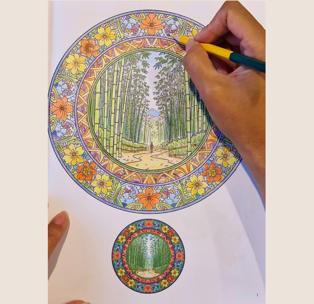 A person is coloring a detailed mandala with floral and bamboo designs. The mandala features a central scene of a bamboo forest pathway with a distant mountain in the background, and it is being filled with various colors using markers.