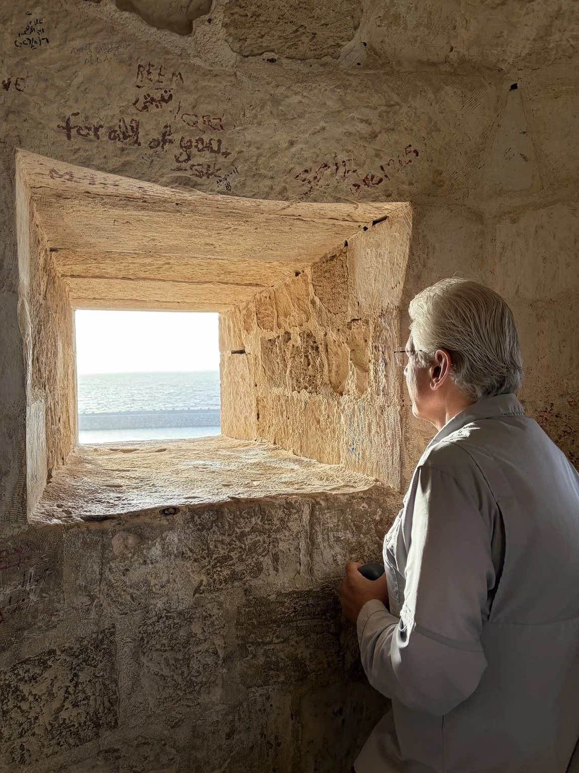 Ali Issa looking through a stone window toward the sea, reflecting the perspective, journey, and quiet moments that shape the work of becoming.