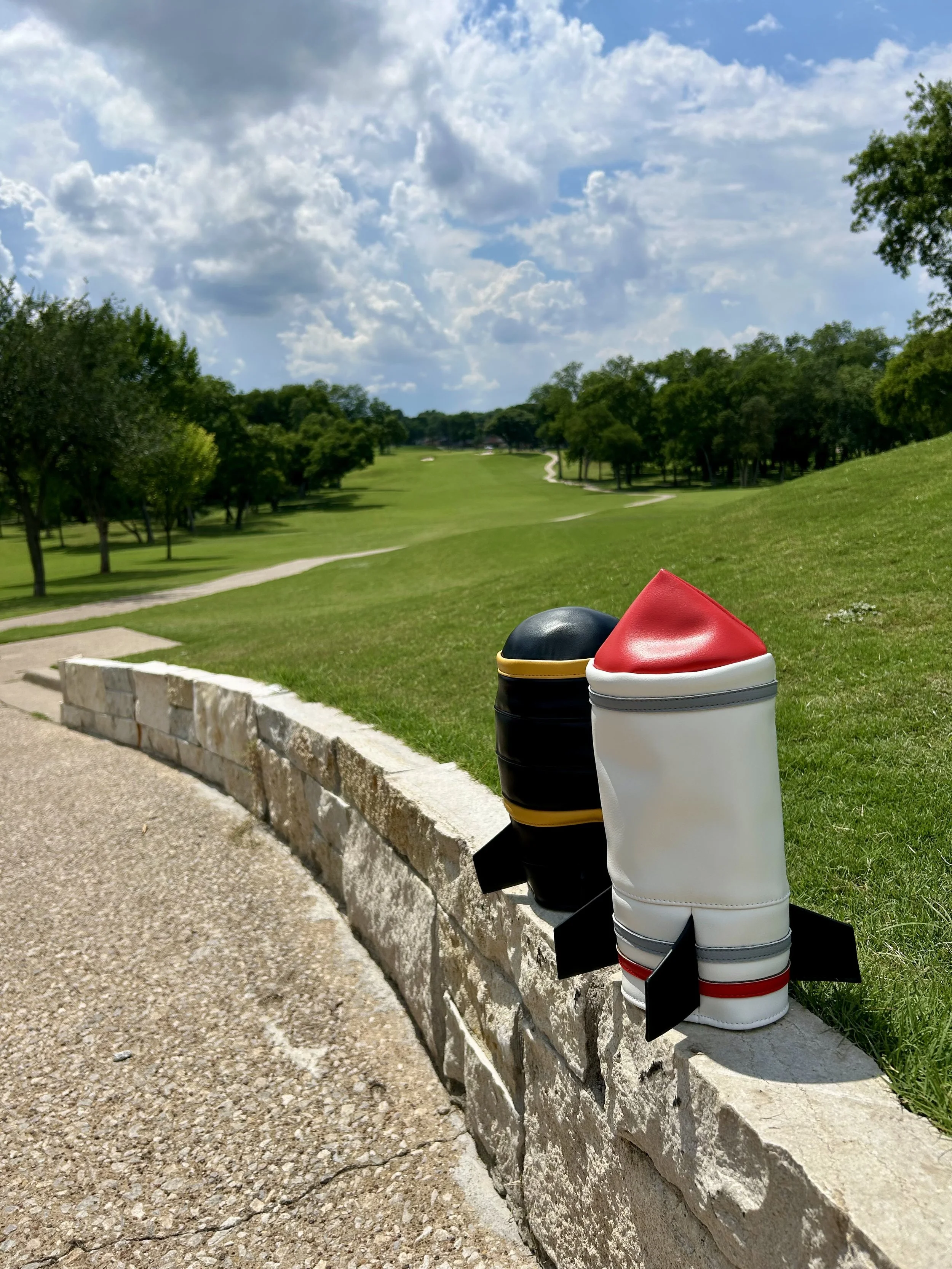 Three golf club head covers, styled to resemble Santa Claus, Santa Elf, and a Black King, are placed on a stone curb at a golf course with green grass, trees, and a partly cloudy sky in the background.