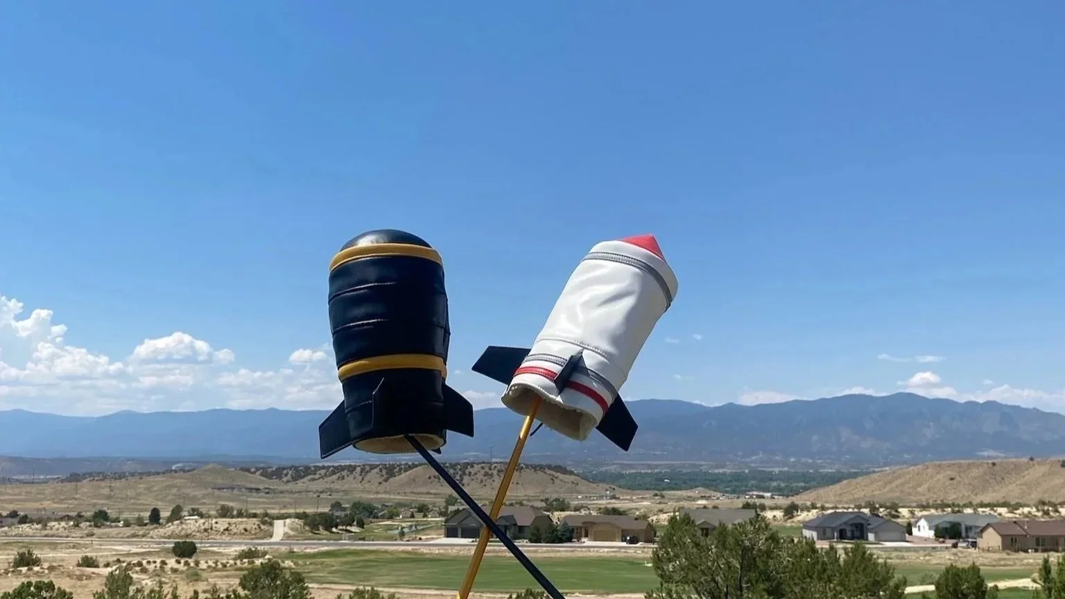 Two small model rockets, one black with yellow stripes and the other white with red and gray accents, mounted on sticks and held upright in an open field with houses and mountains in the background under a blue sky.