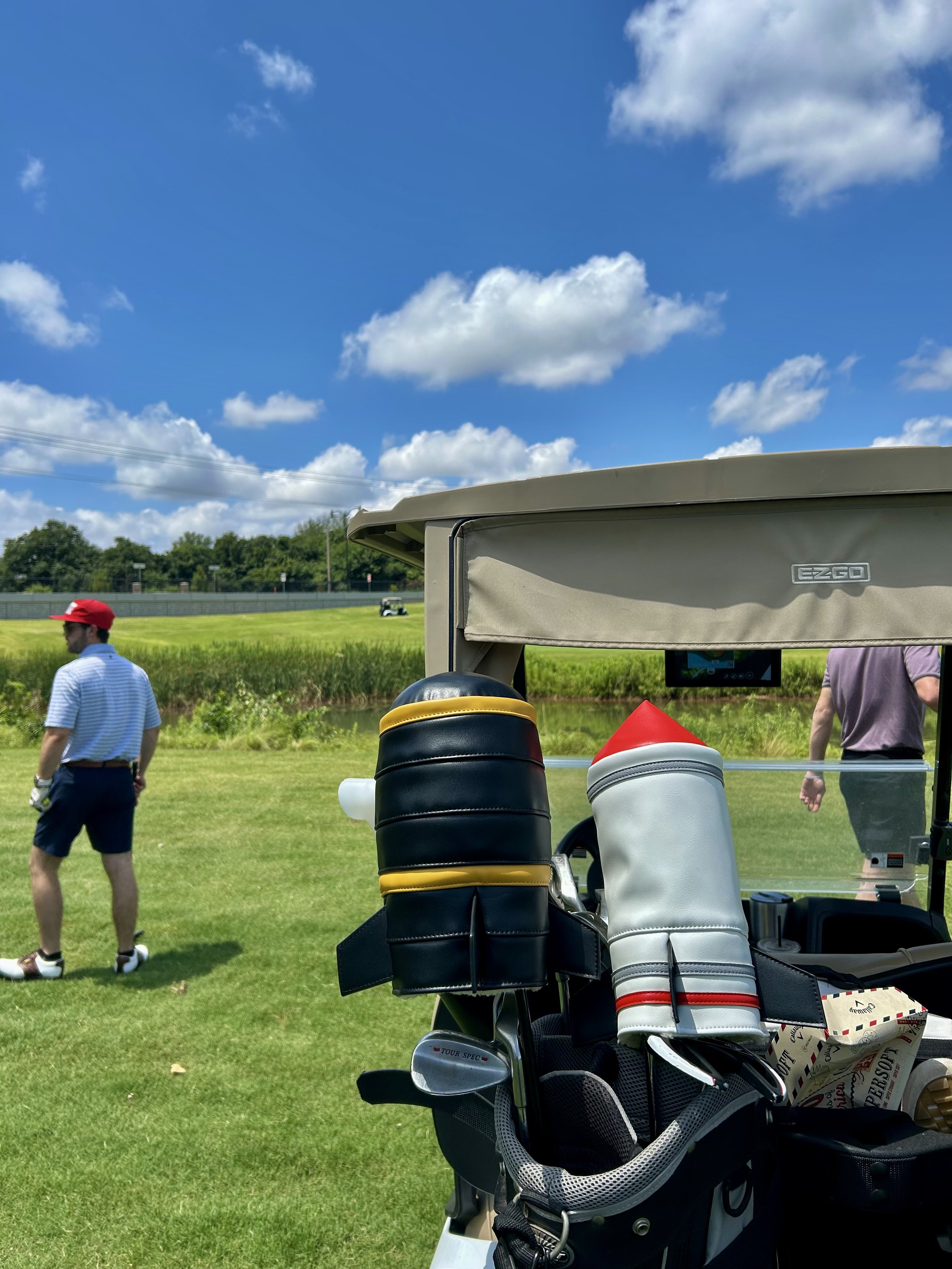 Golf course with golf cart and golf bags in the foreground, two men walking and talking in the middle ground, bright blue sky with scattered clouds in the background.