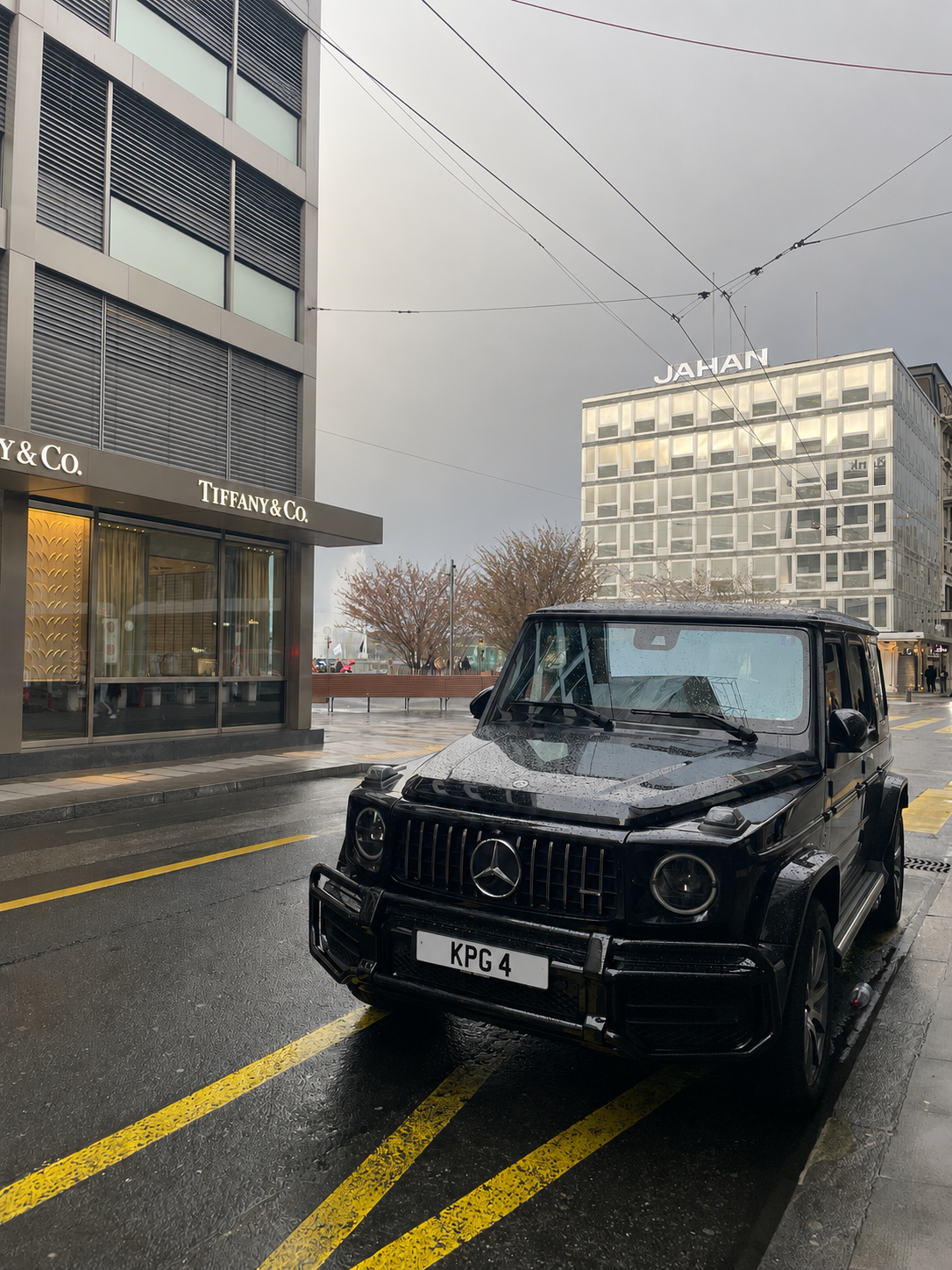 A black Mercedes-Benz G-Class parked on a street in front of a Tiffany & Co. store with a modern building visible in the background and a cloudy sky overhead.