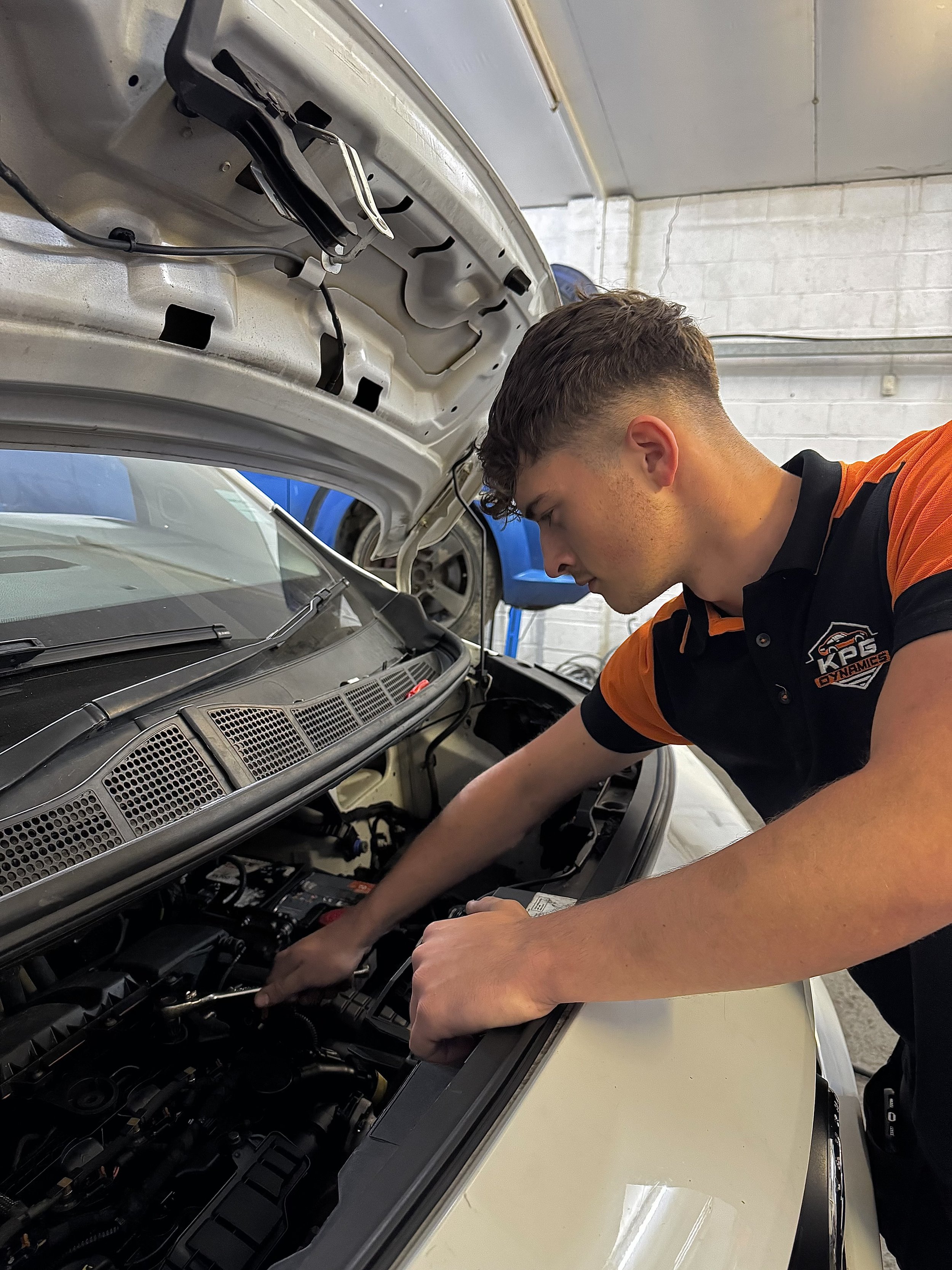 A mechanic wearing an orange and black uniform working under the open hood of a car in a garage.