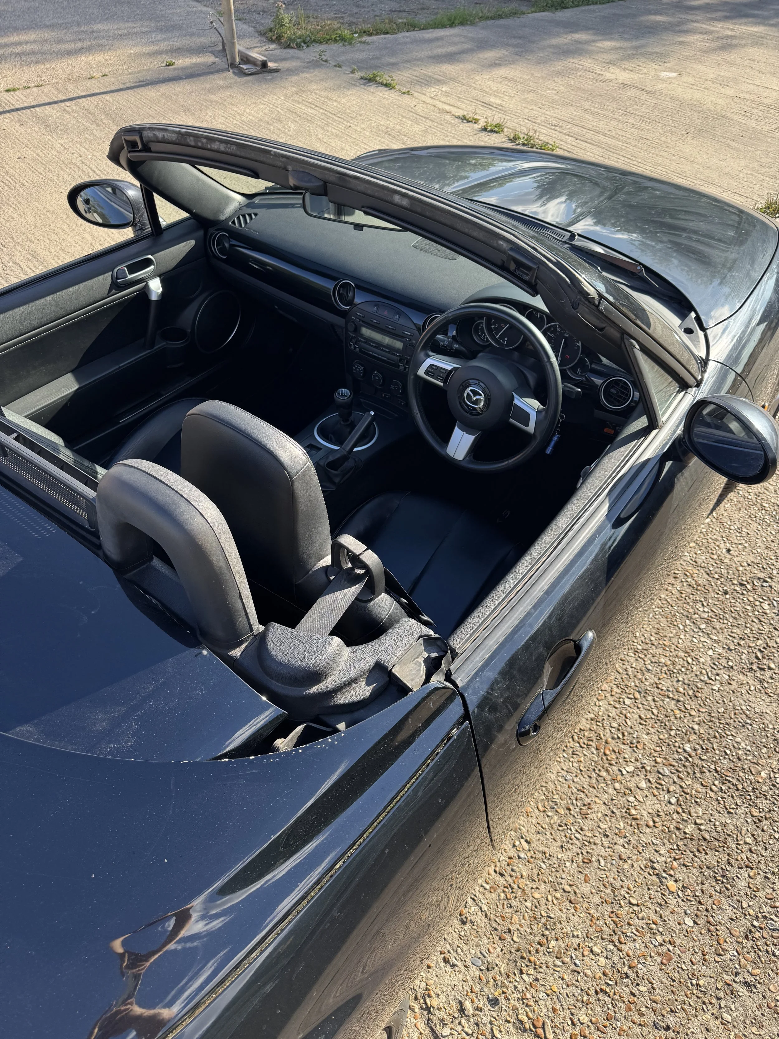 A black Mazda convertible car parked on a gravel and dirt surface, viewed from above, with the top down.