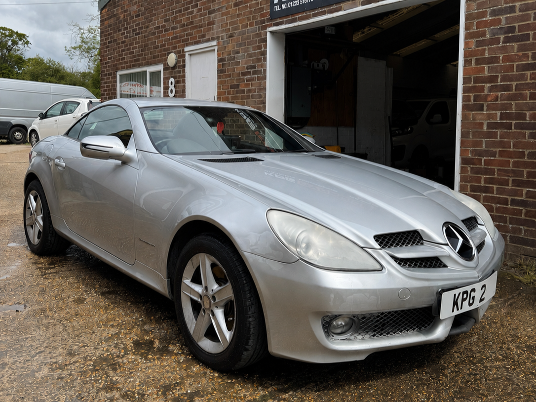 A silver Mercedes-Benz convertible parked outside a brick building with a garage door, a white van and another car visible in the background.