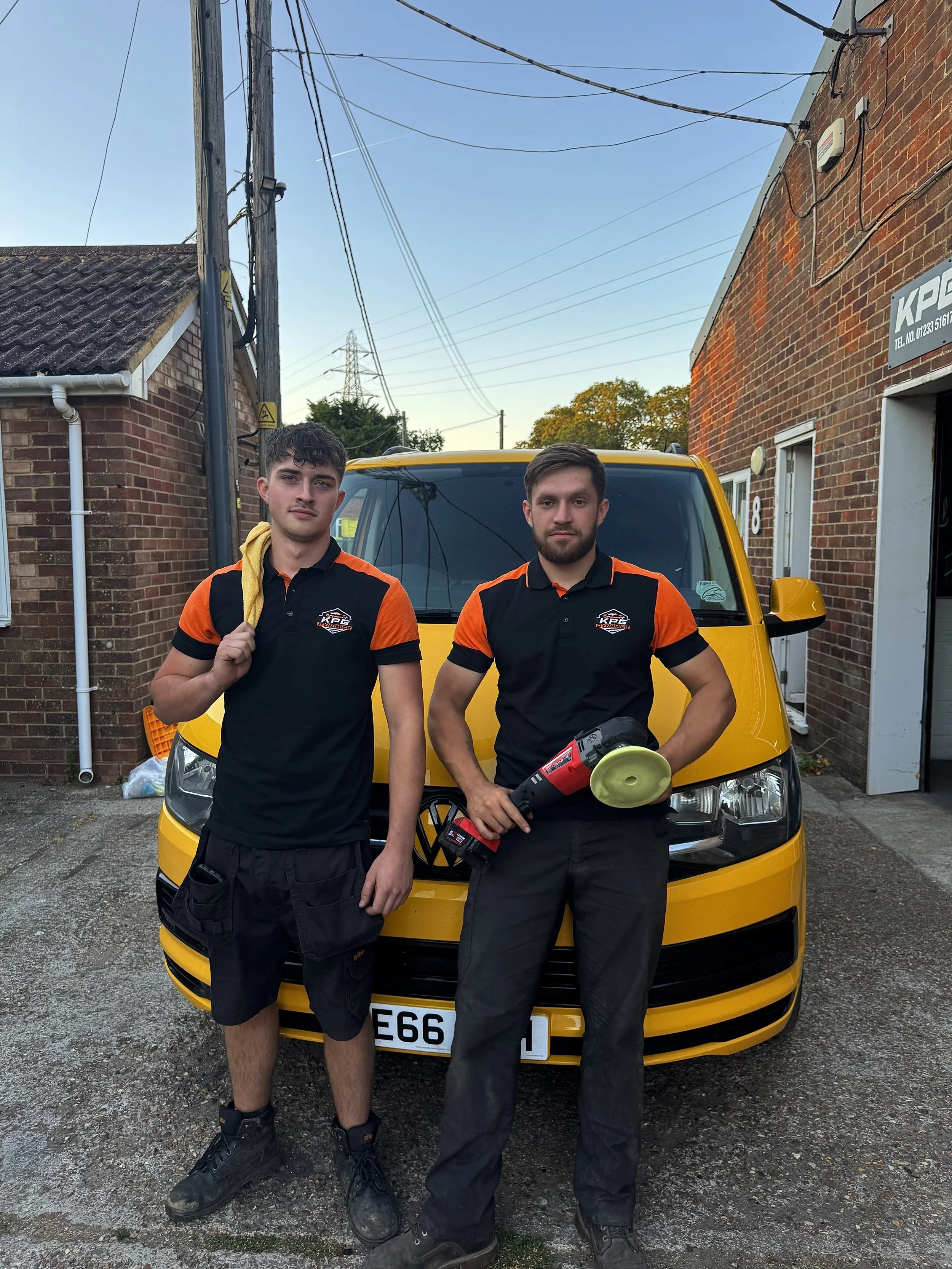 Two young men wearing black and orange uniforms stand in front of a yellow car on a driveway outside brick houses. One man holds a hairdryer, the other holds a megaphone. They are posing for the photo.