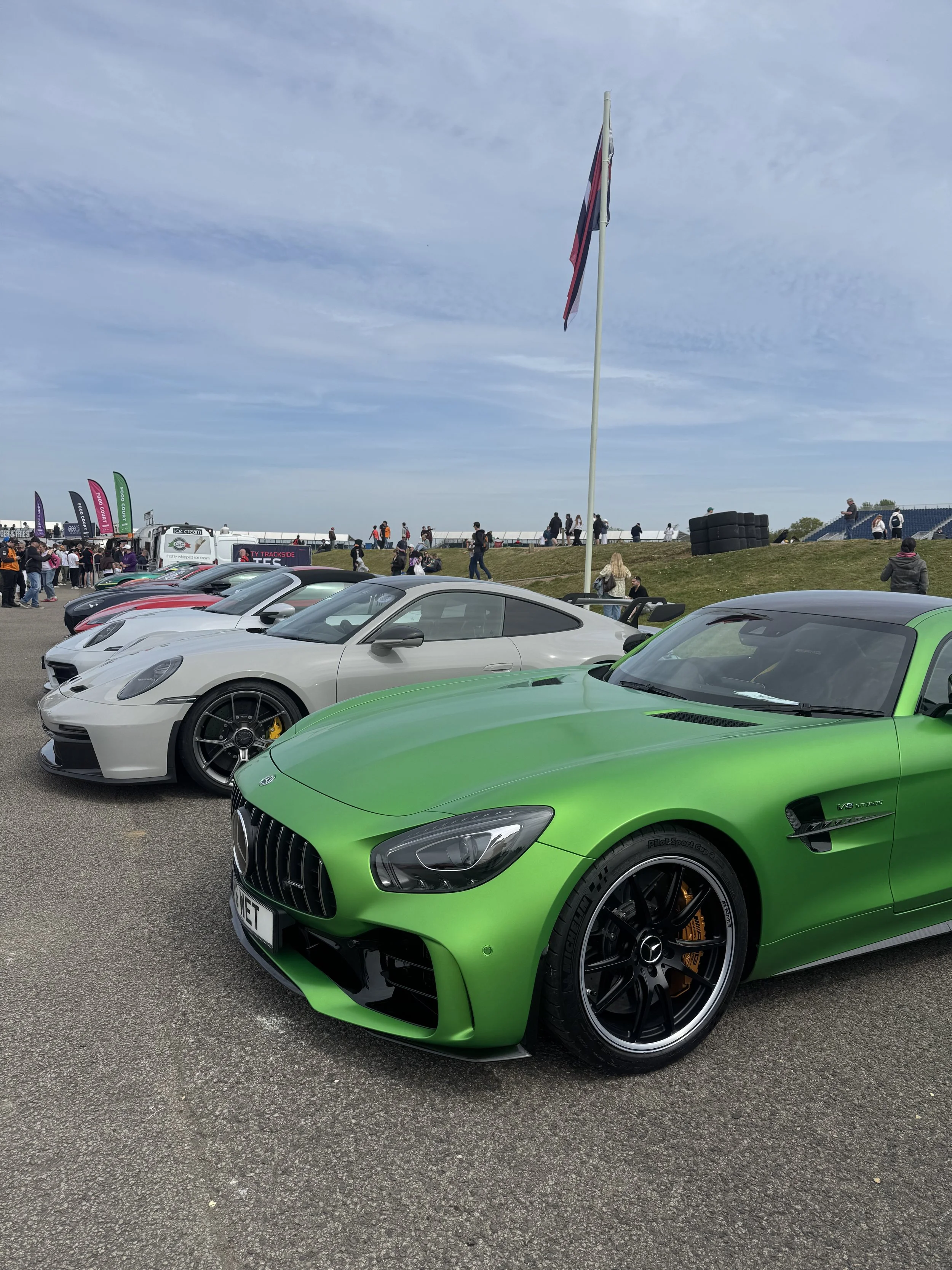 A lineup of luxury cars, including a green car in front, at an outdoor event with people walking and gathering, flags, and a large American flag on a pole in the background under a partly cloudy sky.
