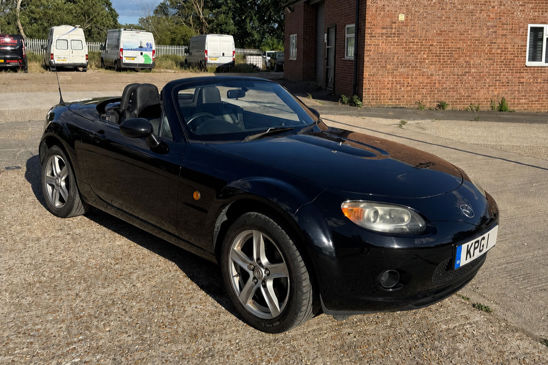 A black Mazda MX-5 Miata convertible parked on a street with brick buildings and white vans in the background.