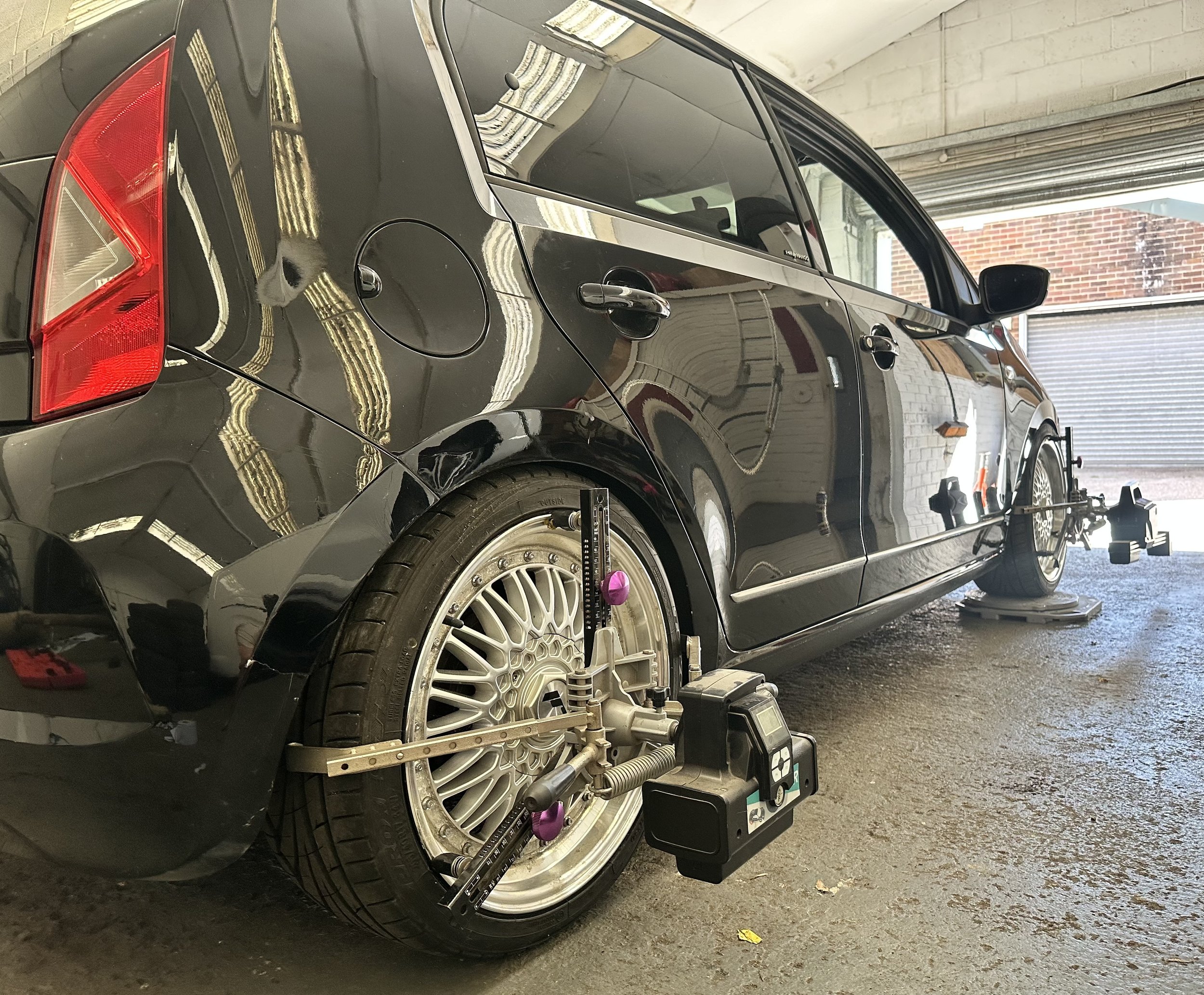 A black car in a garage being serviced, with equipment and tools around it, and a wheel removed, exposing the brake and suspension components.