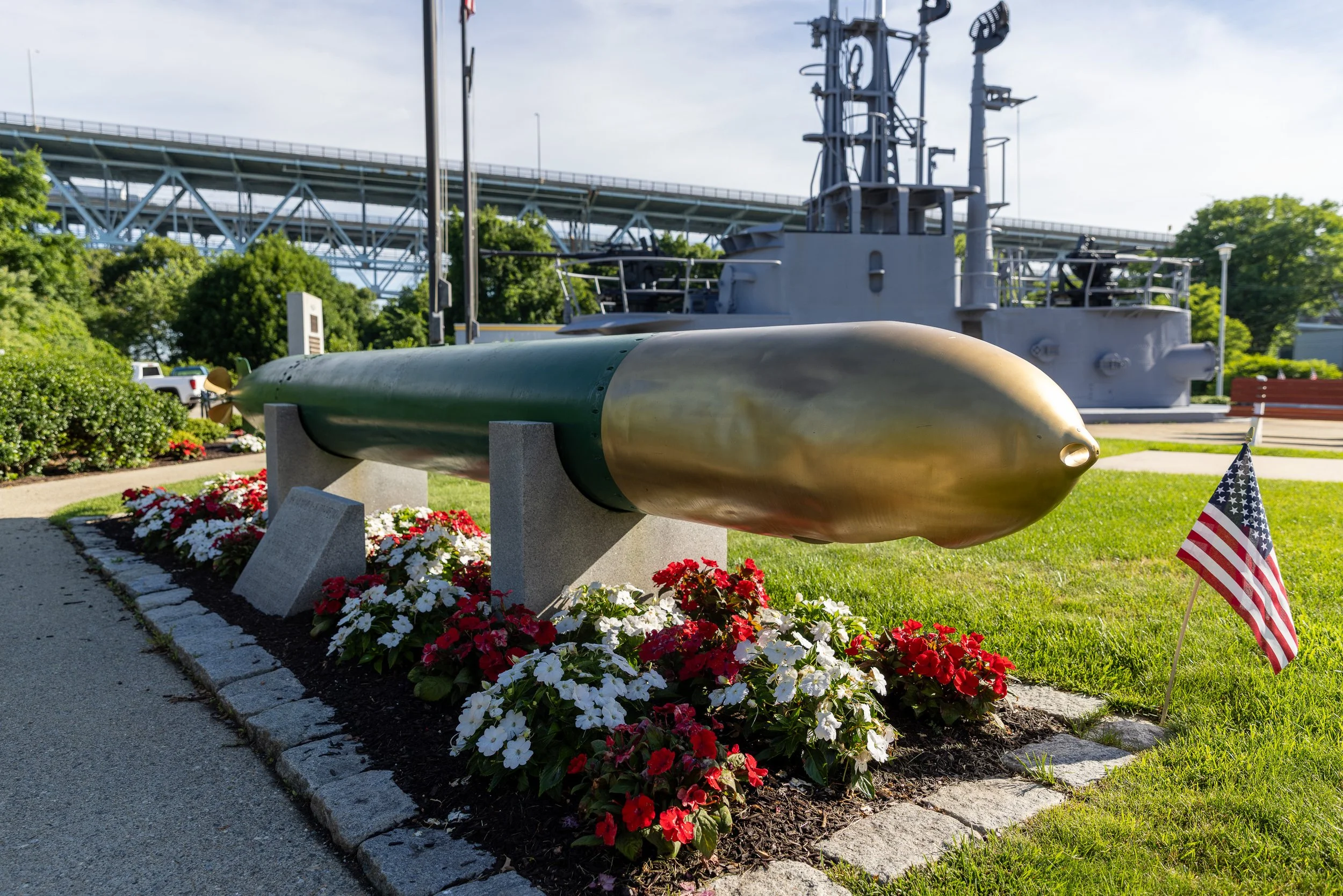 A preserved torpedo fills the foreground at the Submarine Force Museum in Groton, with a historic submarine sail and the Gold Star Memorial Bridge behind it. Weapon, vessel, river, and highway converge in a single view of the town that became the Sub