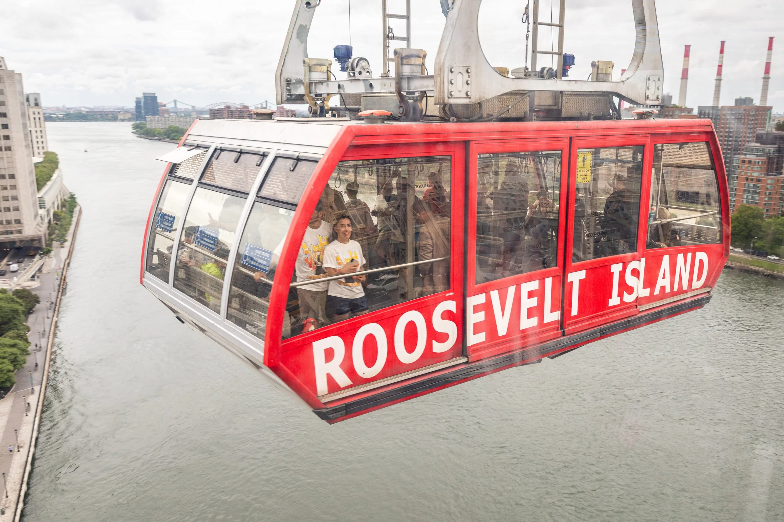 Red Roosevelt Island Tramway cabin carrying passengers above the East River, with New York City buildings in the background.