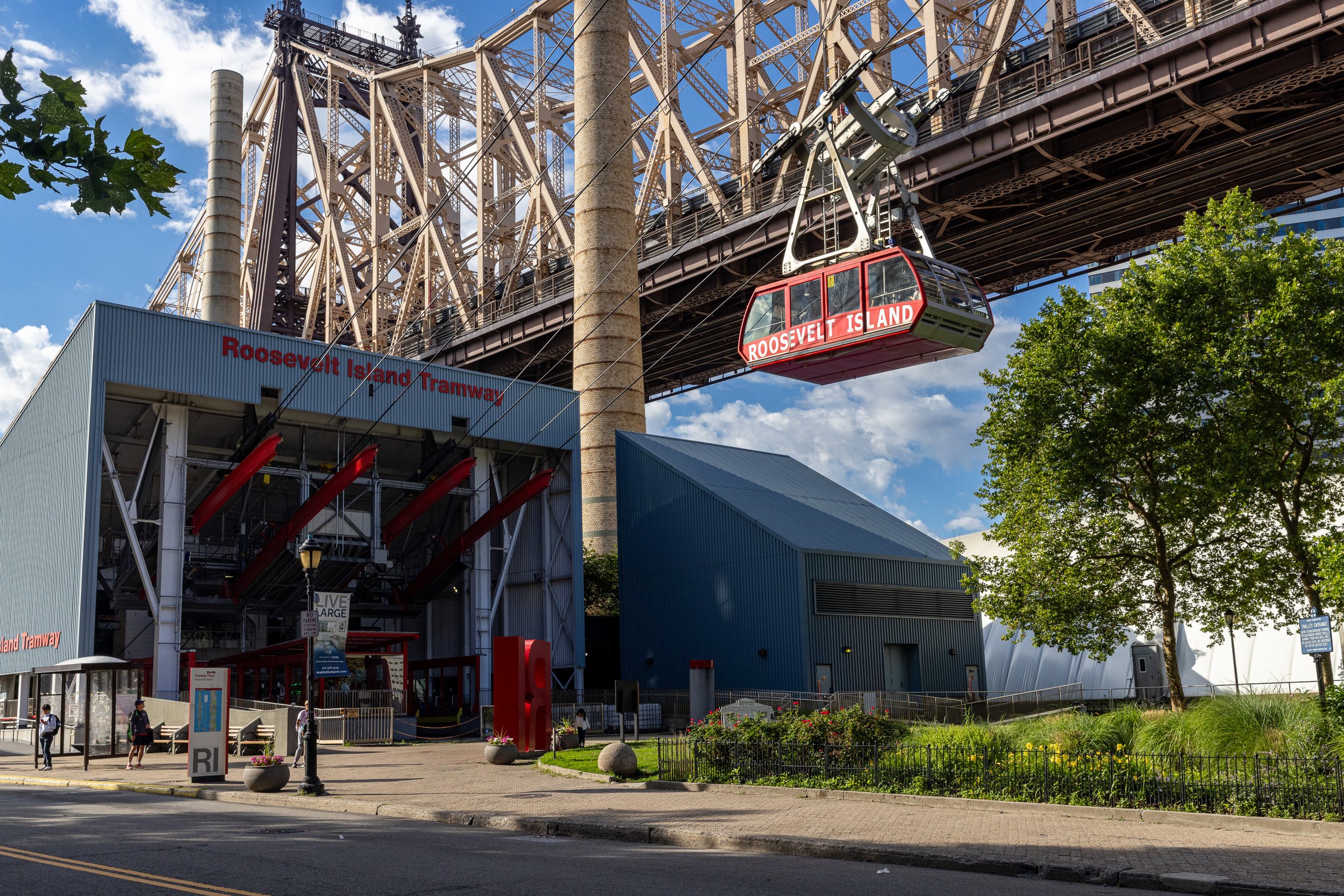 The Roosevelt Island Tramway station sits beneath the Queensboro Bridge as a red tram cabin travels beside it.