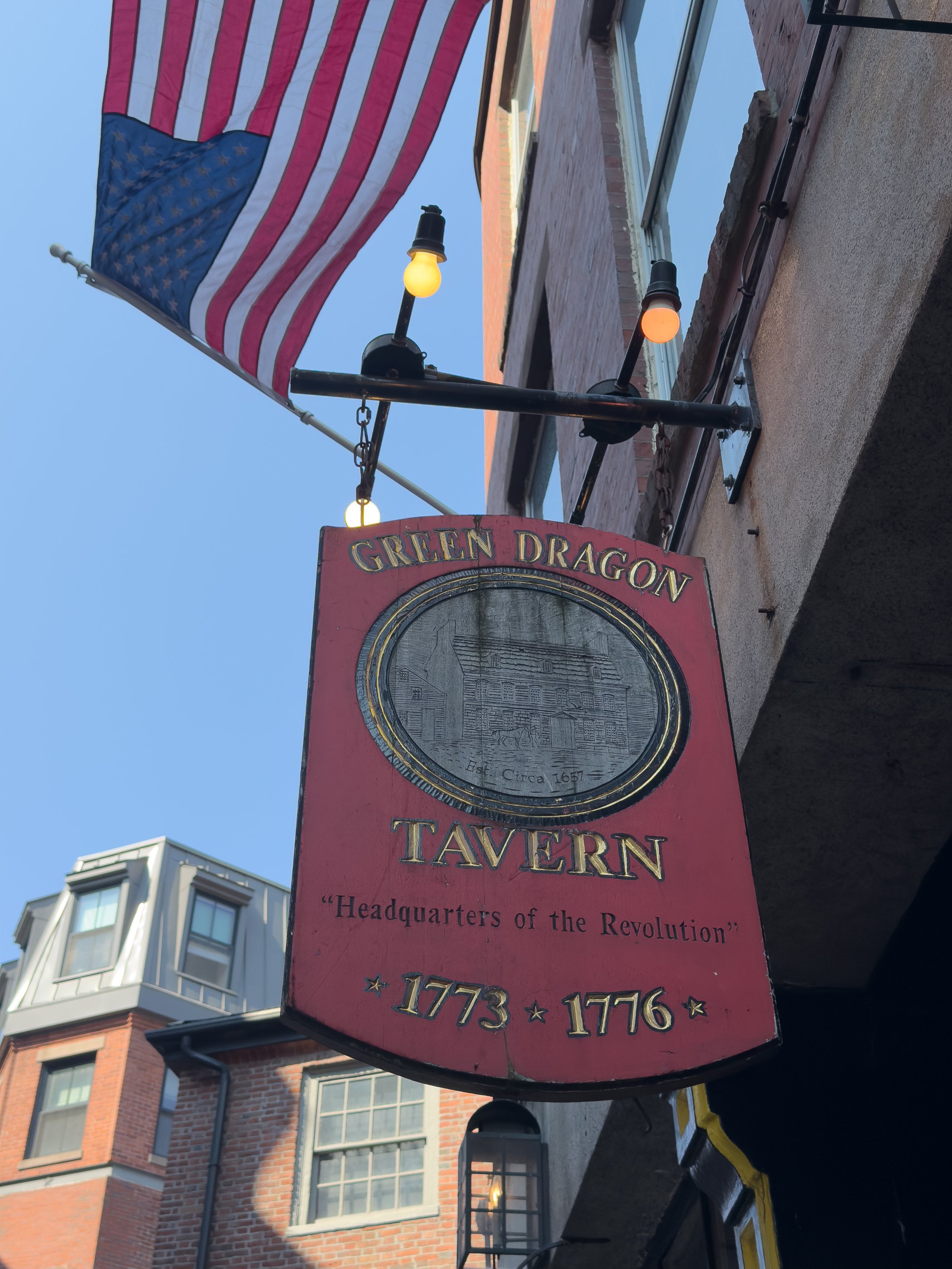 Red Green Dragon Tavern sign beneath an American flag and glowing bulbs on a narrow Boston street.