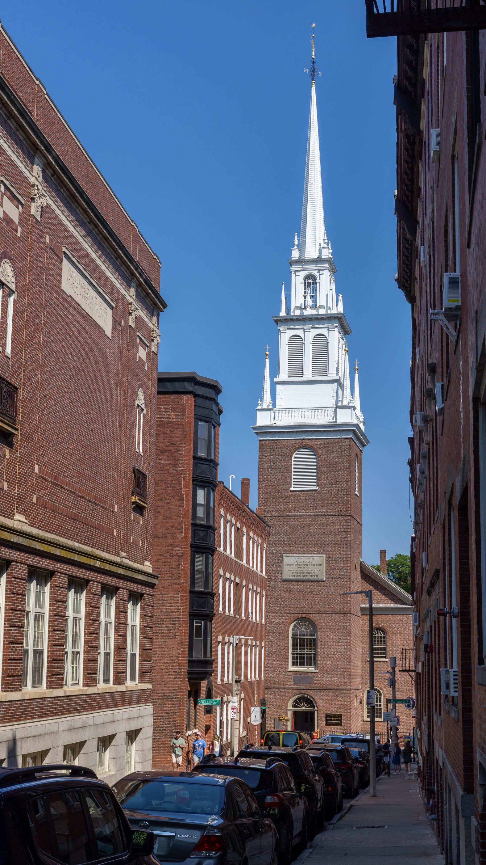 Old North Church framed by brick buildings and parked cars along narrow Hull Street in Boston’s North End.