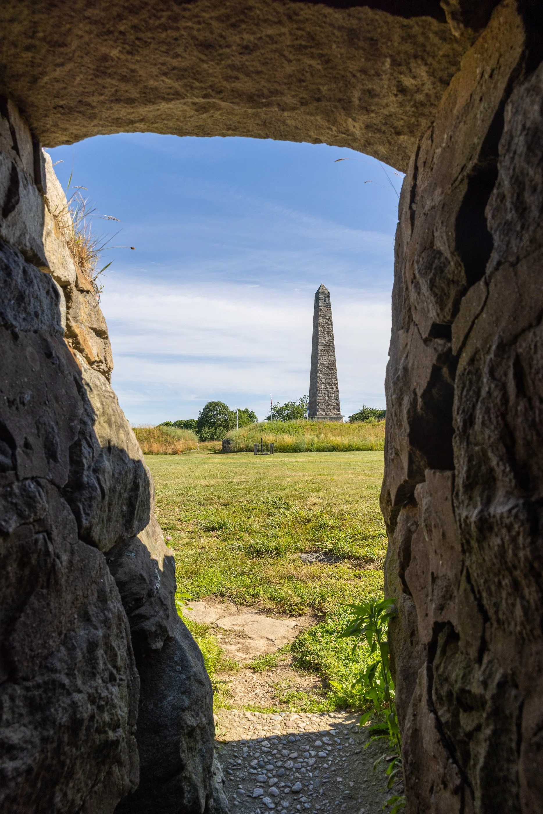 A stone passage through Fort Griswold’s earthworks frames the Groton Monument beyond it. The view links the surviving Revolutionary War fort with the memorial built to honor the defenders who died there.