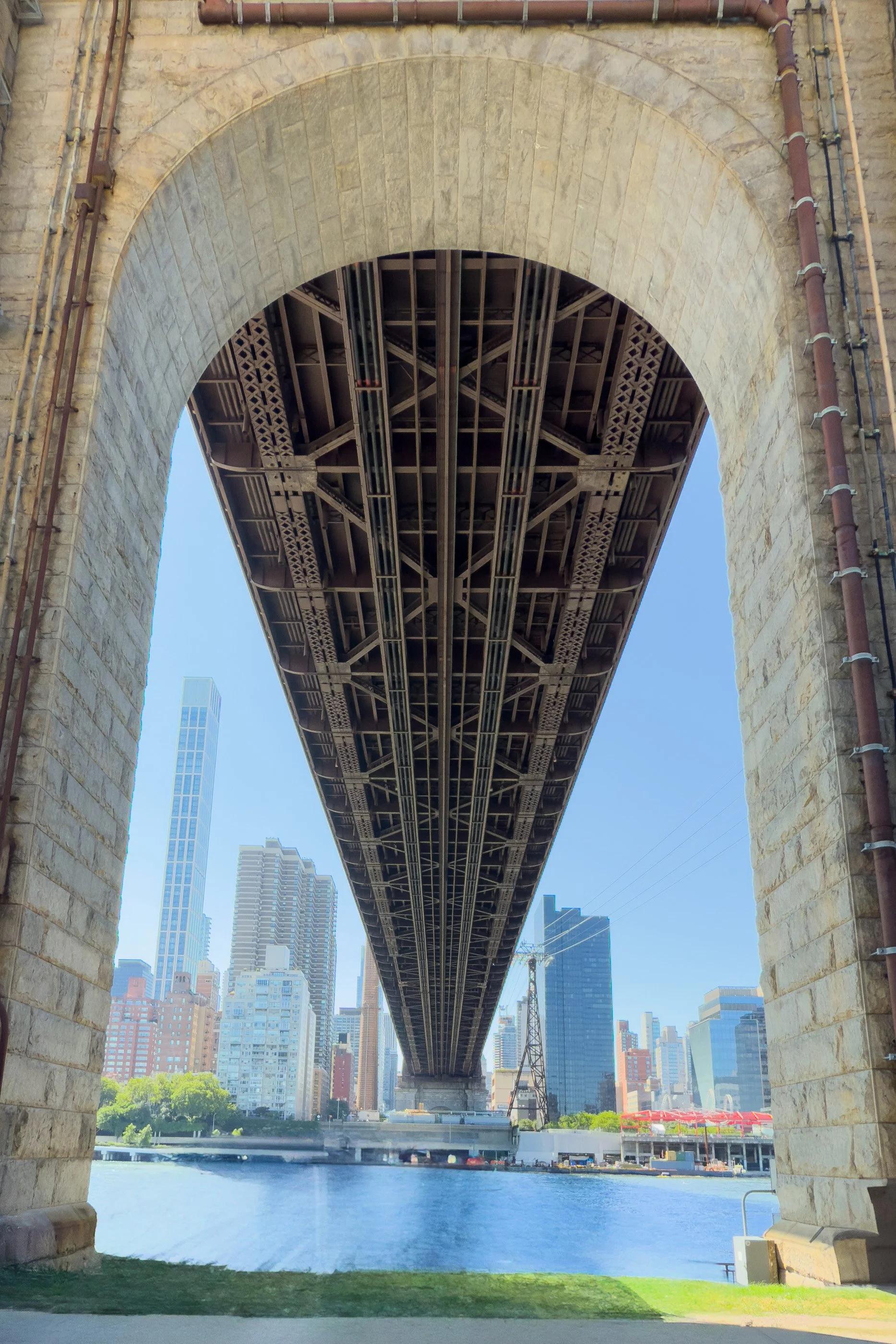 The underside of the Queensboro Bridge fills the view above Roosevelt Island, with the East River and city buildings beyond.
