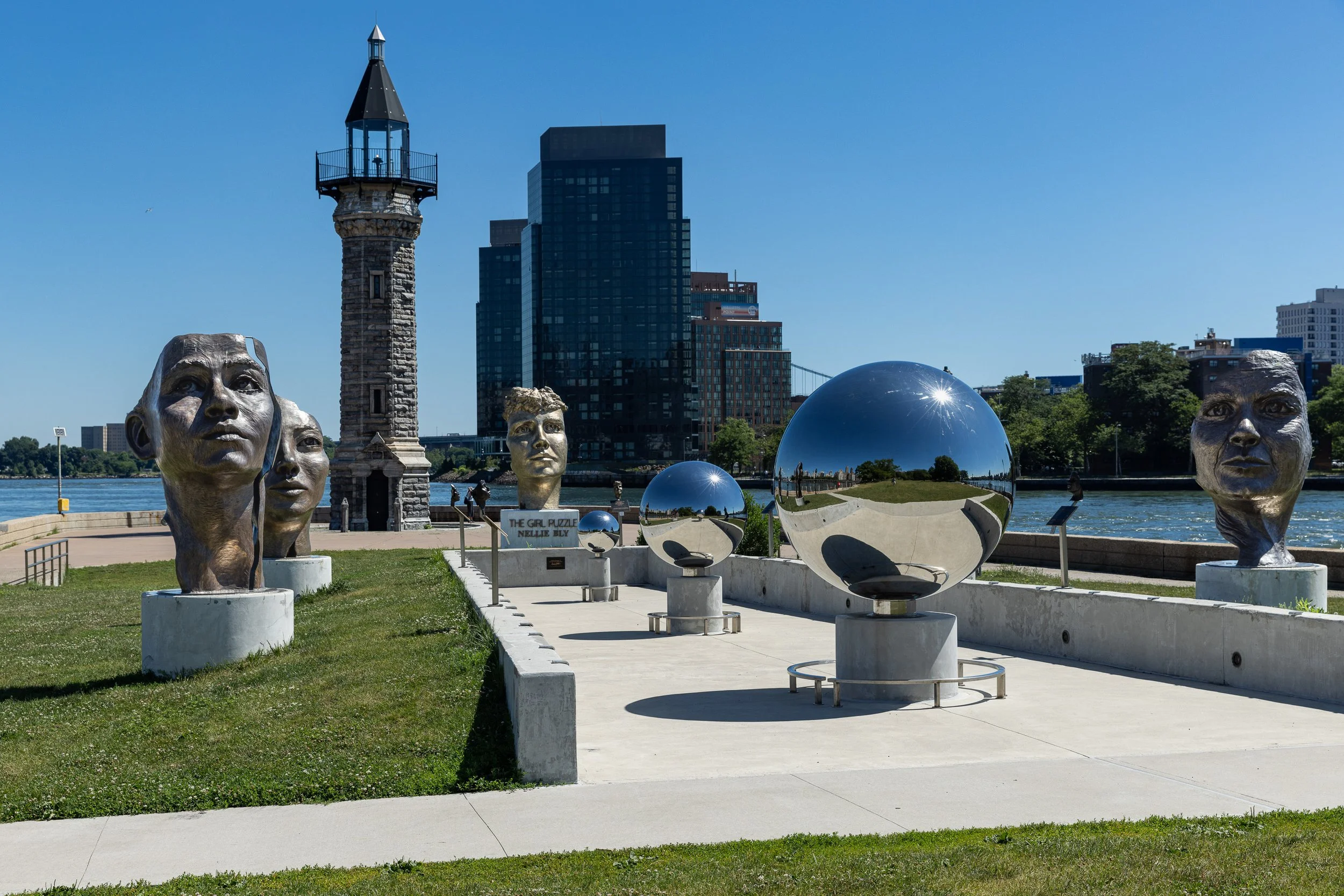 Bronze faces and mirrored spheres of The Girl Puzzle sculpture stand on a lawn with the Roosevelt Island Lighthouse behind them.