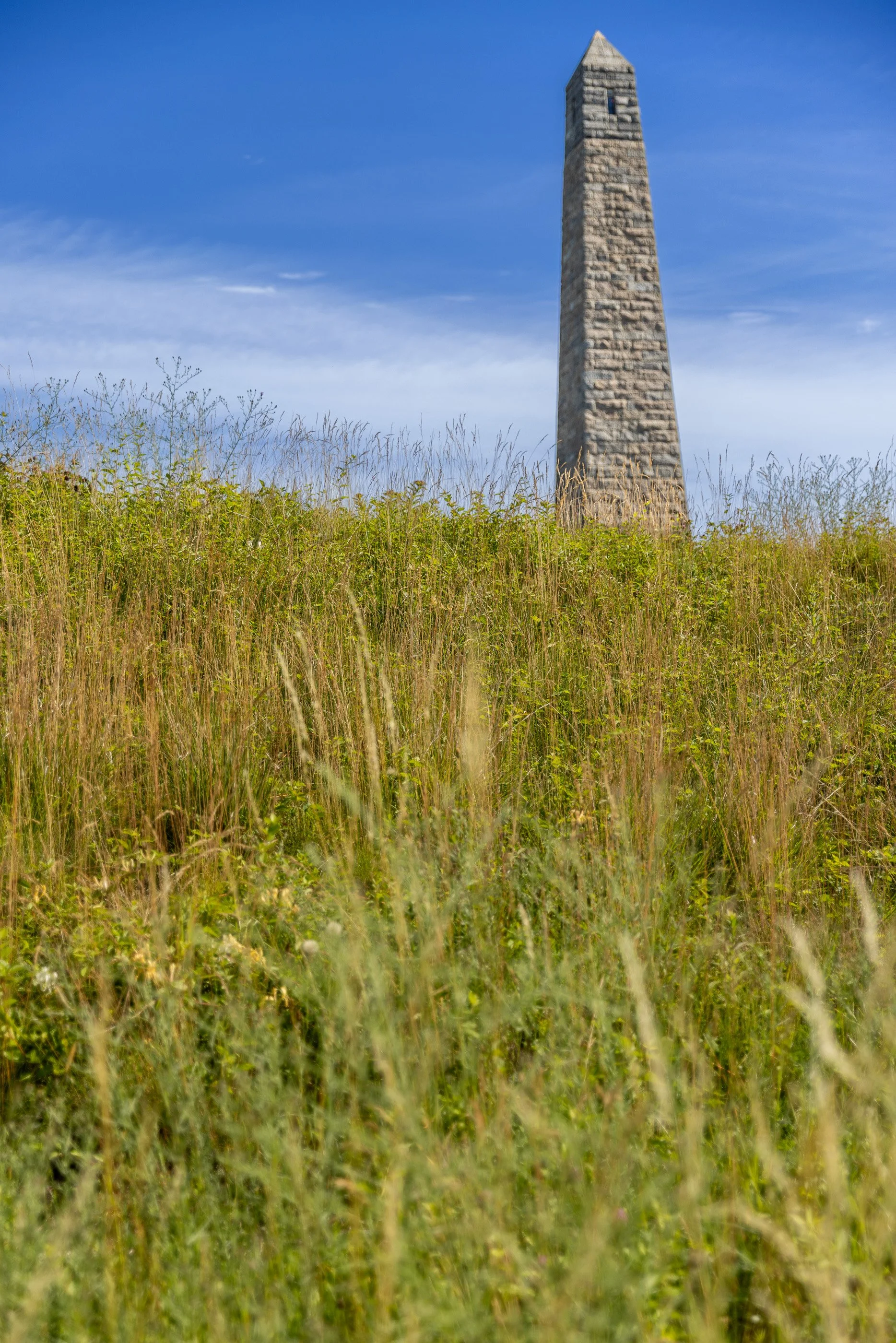 The Groton Monument rises beyond the tall grass and surviving earthworks of Fort Griswold. From this angle, the memorial’s direct relationship to the battlefield it commemorates becomes impossible to miss.