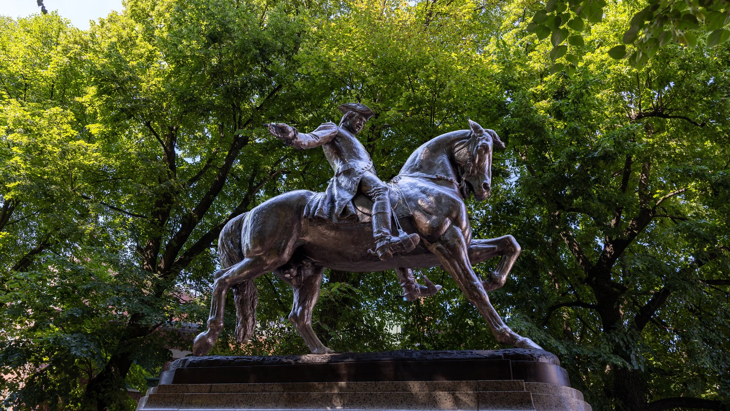 Side view of Cyrus Dallin’s bronze Paul Revere monument, with the horse and rider framed by green trees.