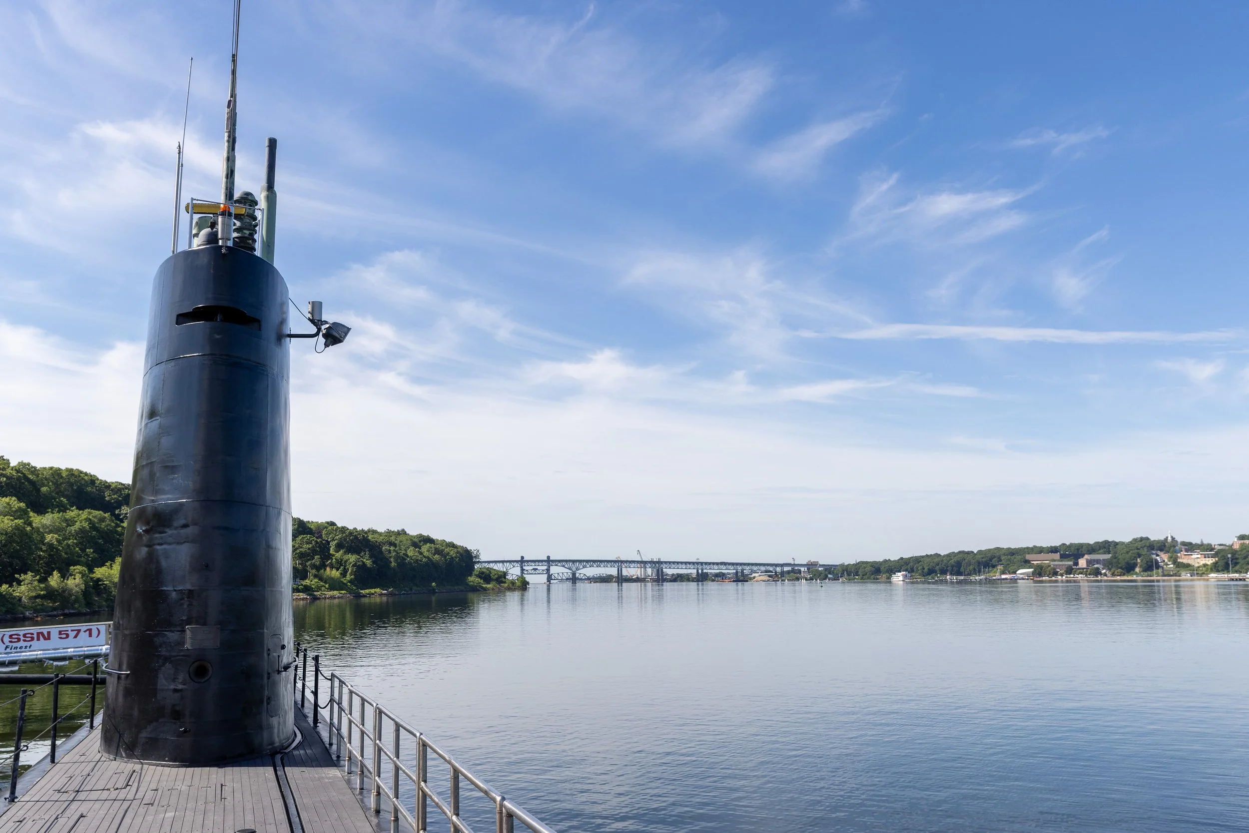 The sail of USS Nautilus stands beside a broad view up the Thames River toward the Gold Star Memorial Bridge. Built at Electric Boat in Groton, the submarine remains inseparable from the river where she was launched and first headed to sea.