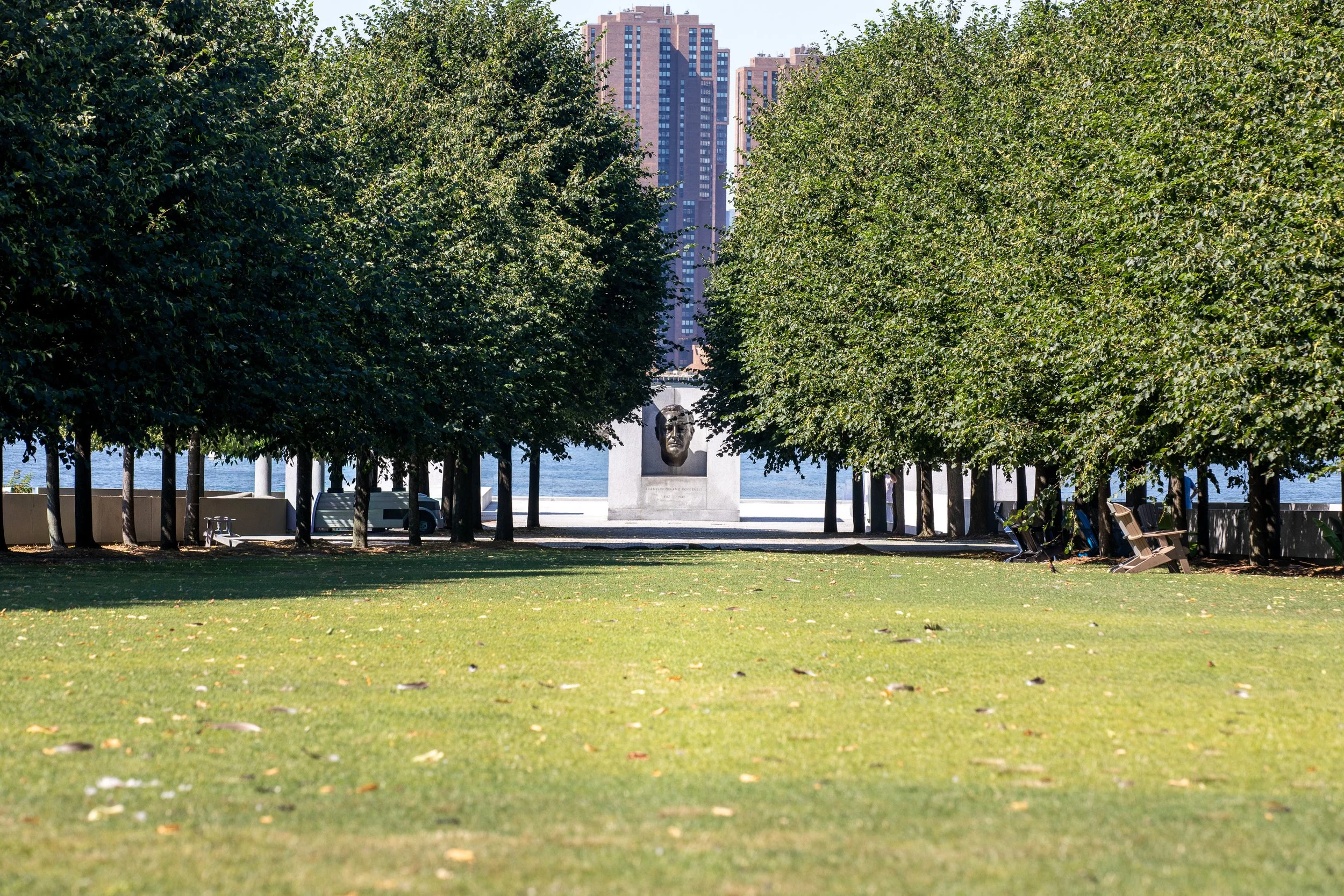 Rows of trees frame the lawn at Franklin D. Roosevelt Four Freedoms Park, with Roosevelt’s bust at the far end and city buildings beyond.