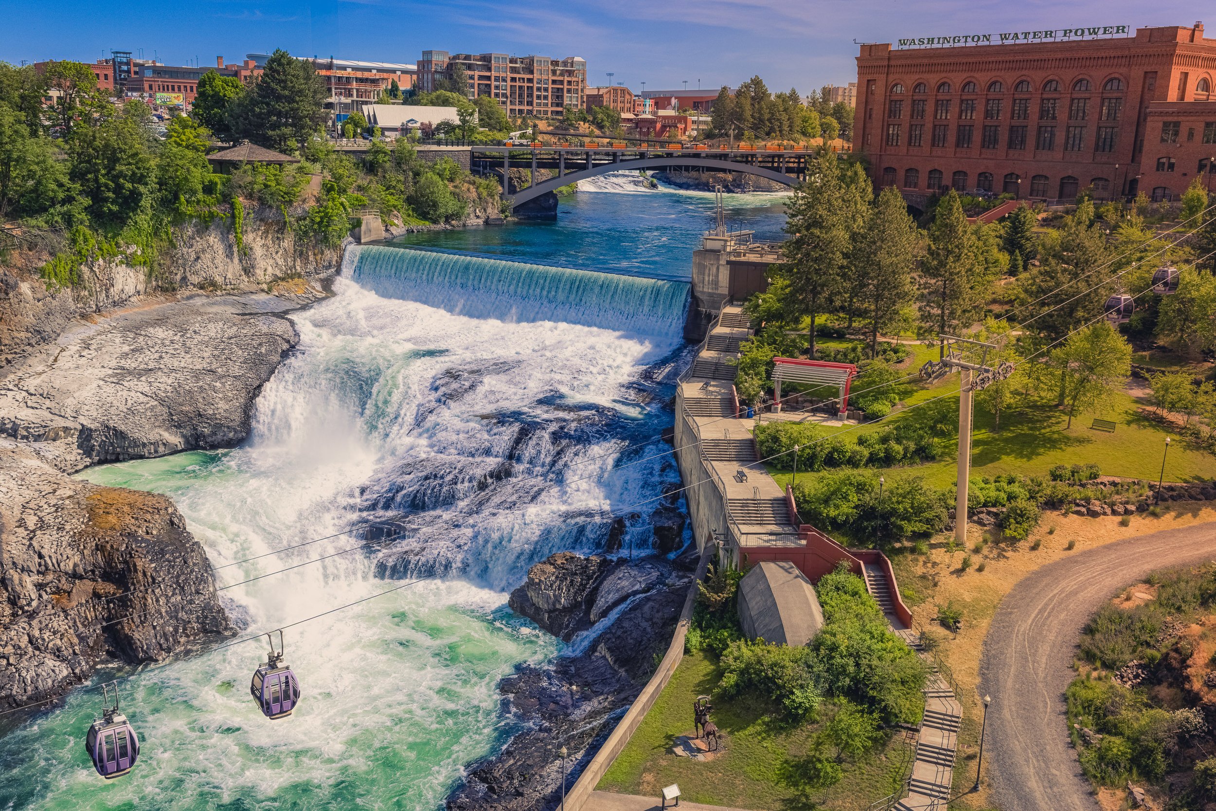 SkyRide gondolas pass over Lower Spokane Falls below Monroe Street Dam, with the Post Street Substation and Post Street Bridge upstream. The scene brings recreation, transportation, and electrical infrastructure together in a single downtown view.