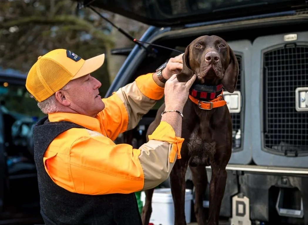A man wearing a yellow hat and orange uniform holding a brown dog, possibly a hunting or working dog, in front of a vehicle with a dog crate.