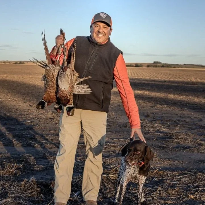 A man smiling and holding a bird he hunted, standing in a field with a dog by his side.