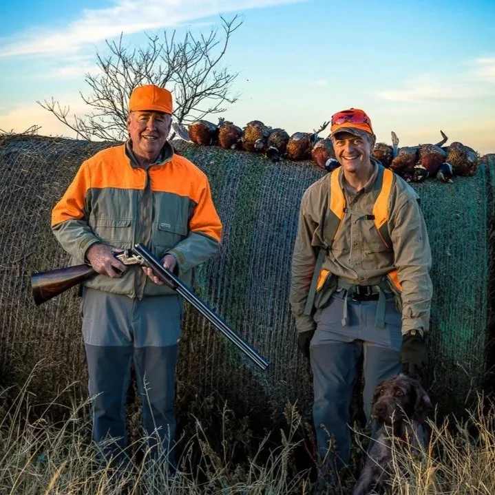 Two hunters, one holding a shotgun, standing outdoors with a row of pheasants hanging behind them, and a hunting dog sitting nearby.