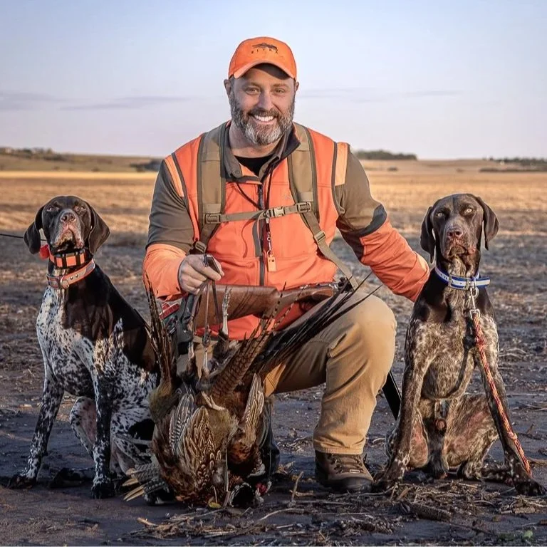 A man in orange hunting gear kneels outdoors with two hunting dogs and a bird at his feet, in an open field.